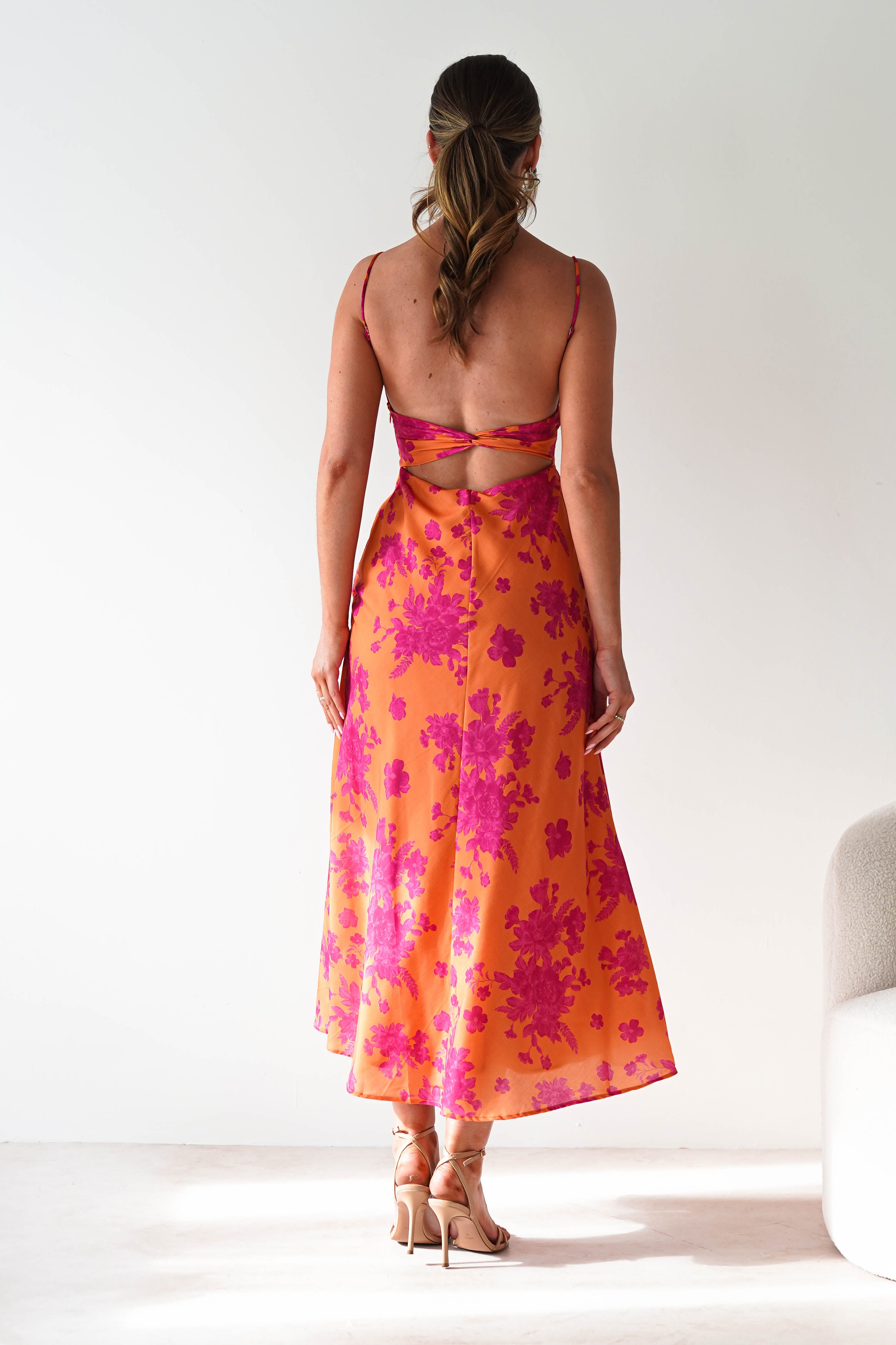 Woman standing with her back to the camera, wearing a sleeveless, orange and pink floral wedding guest dress with an open back, spaghetti strap details, and nude high-heeled sandals. She is in a minimal, light-colored room.