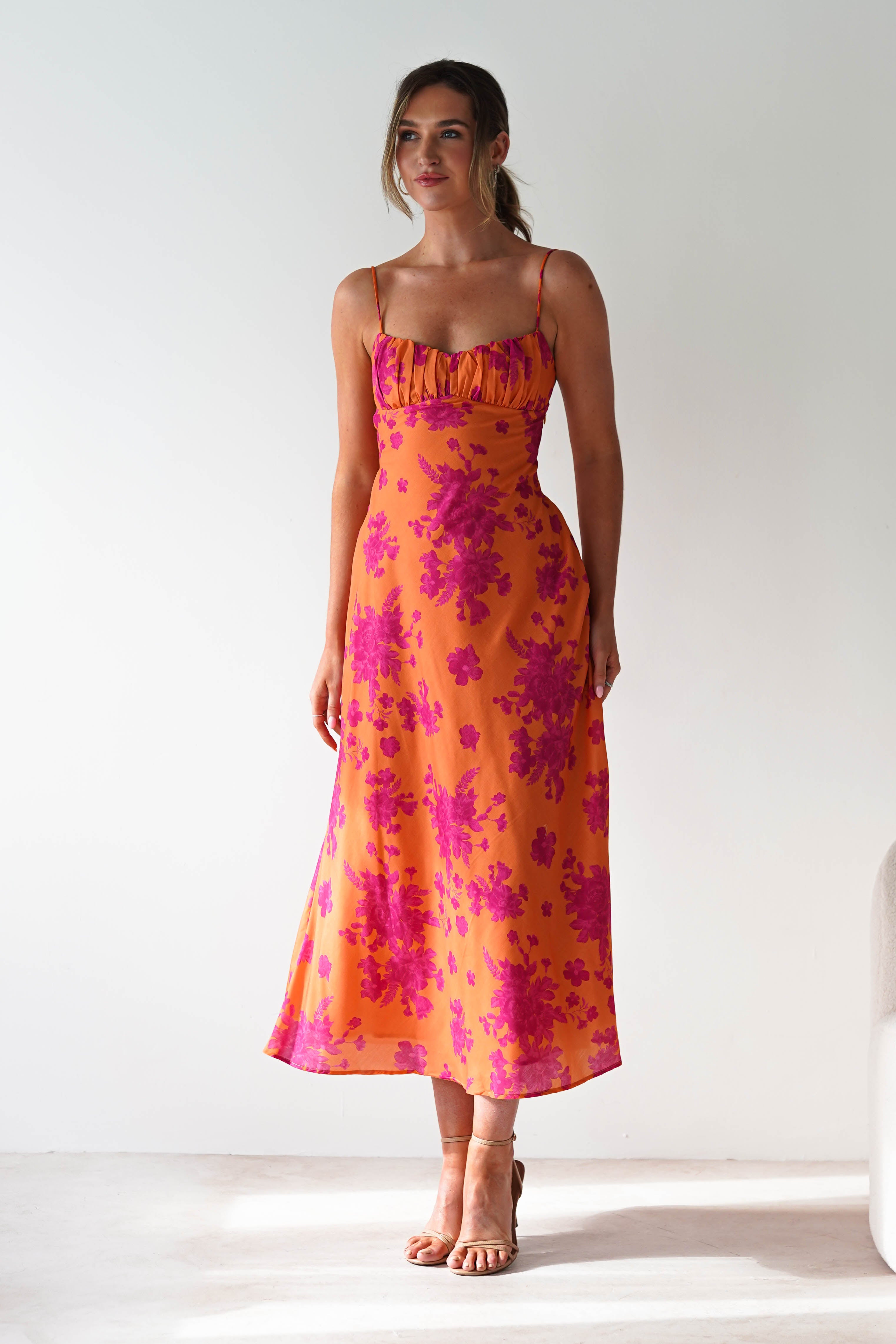 A woman stands in a brightly lit room wearing an orange wedding guest dress with pink floral patterns, thin spaghetti straps, and tan heeled sandals. She has light brown hair tied back and looks slightly to the side.