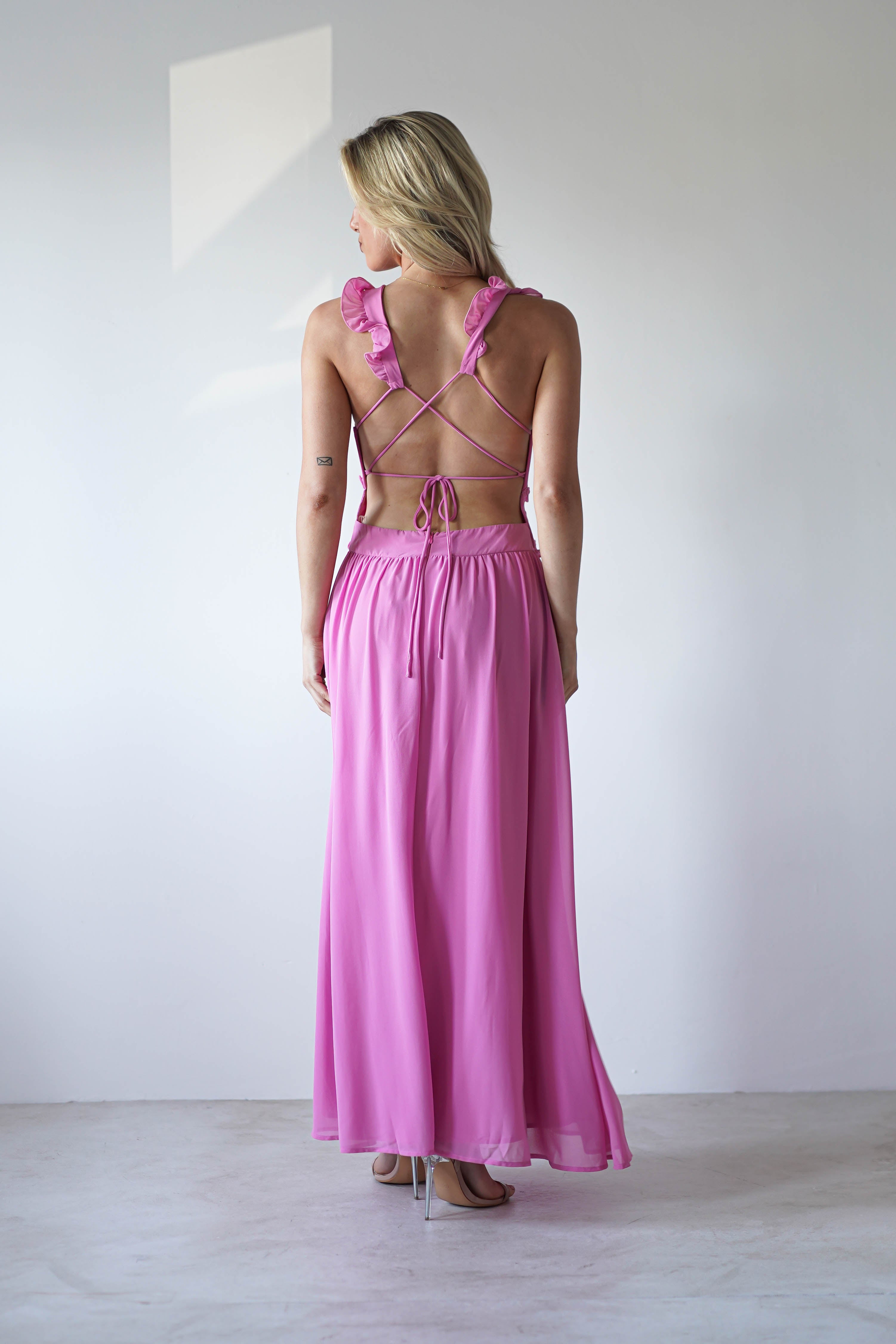 Woman standing in a minimal studio wearing a long, flowing chiffon maxi pink dress with an open back and crisscross straps, perfect for special occasions or as a wedding guest look, facing away from the camera.