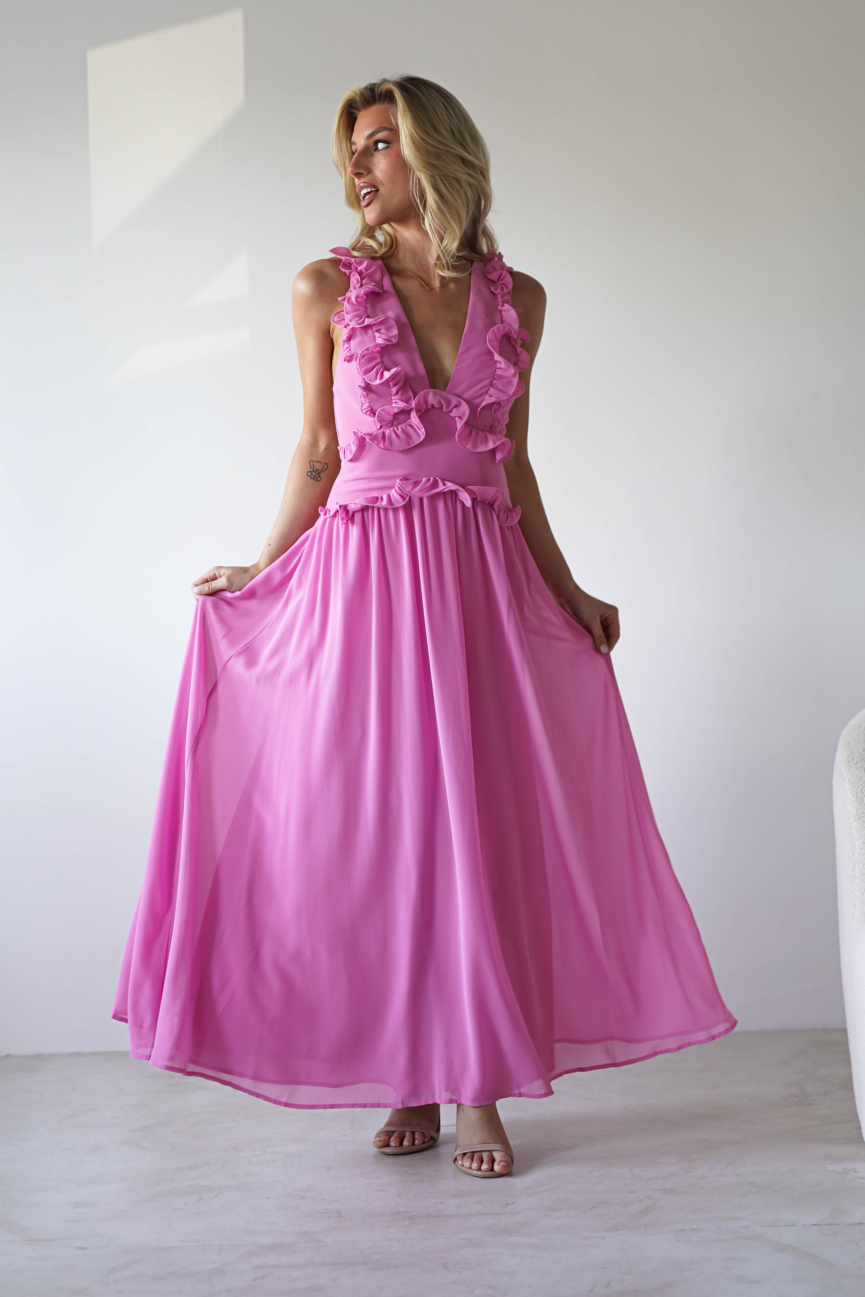 A woman in a sleeveless, floor-length bright pink chiffon maxi dress with ruffled details stands indoors, holding out her skirt. Perfect for wedding guest looks or special occasions, the minimal background features white walls and a light chair.