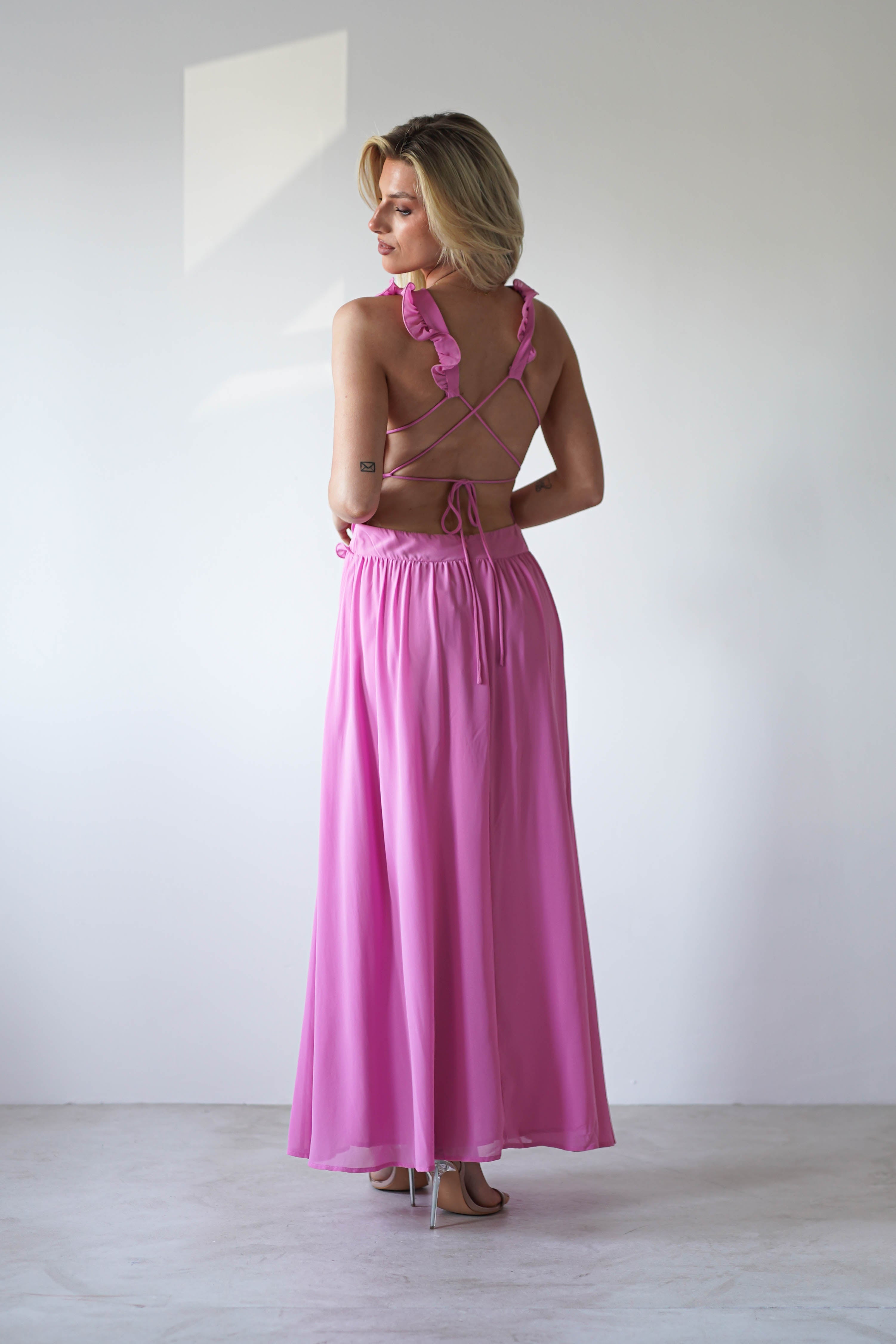 A woman stands facing away in a chiffon maxi pink dress with crisscross straps and ruffle details—an elegant choice for wedding guest style or special occasions, set against a plain white background.
