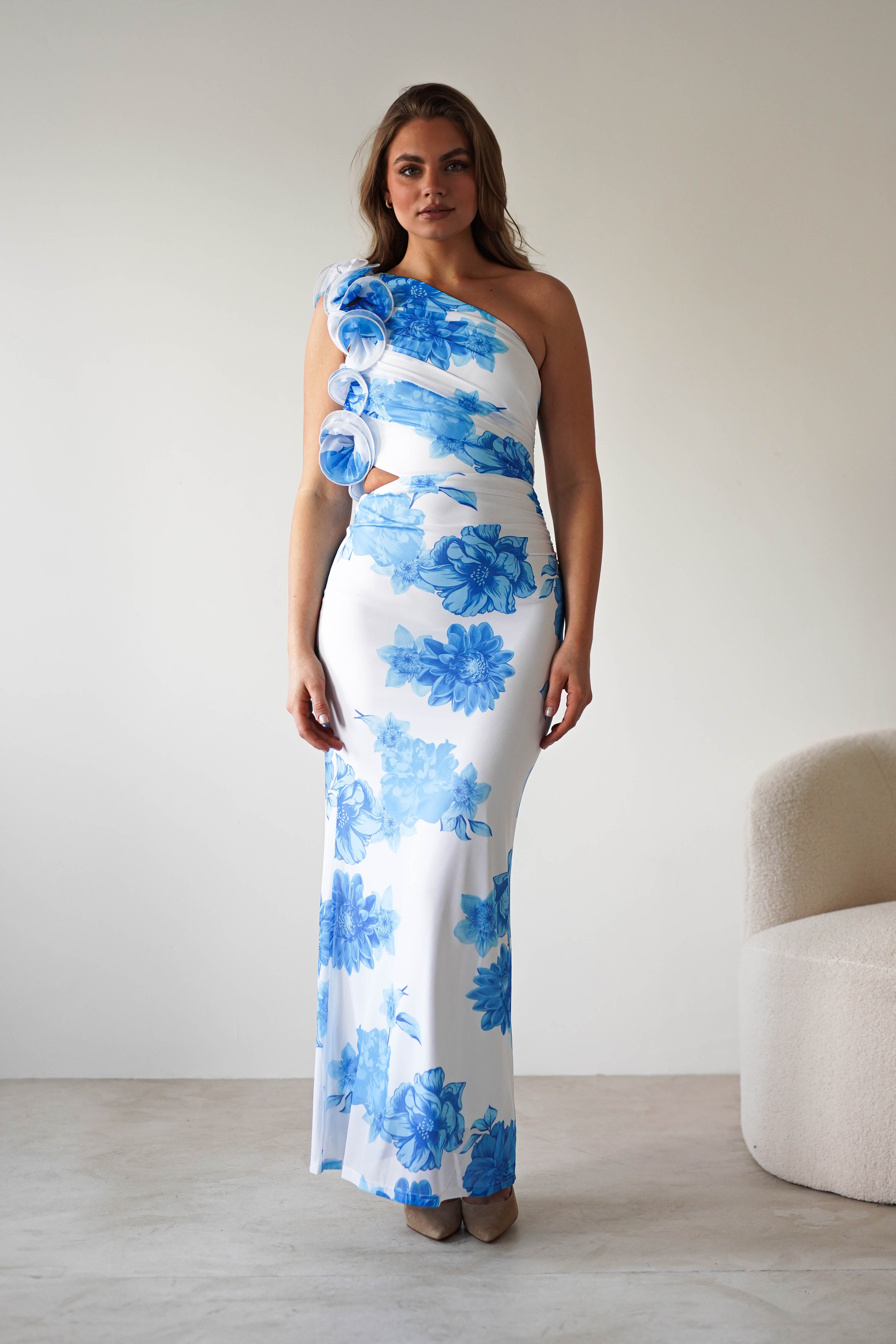 A woman stands in a minimalistic room wearing a long, white one-shoulder wedding guest dress with summer blue print and 3D floral ruffle feature on the shoulder. She poses confidently beside a light-colored, modern chair.