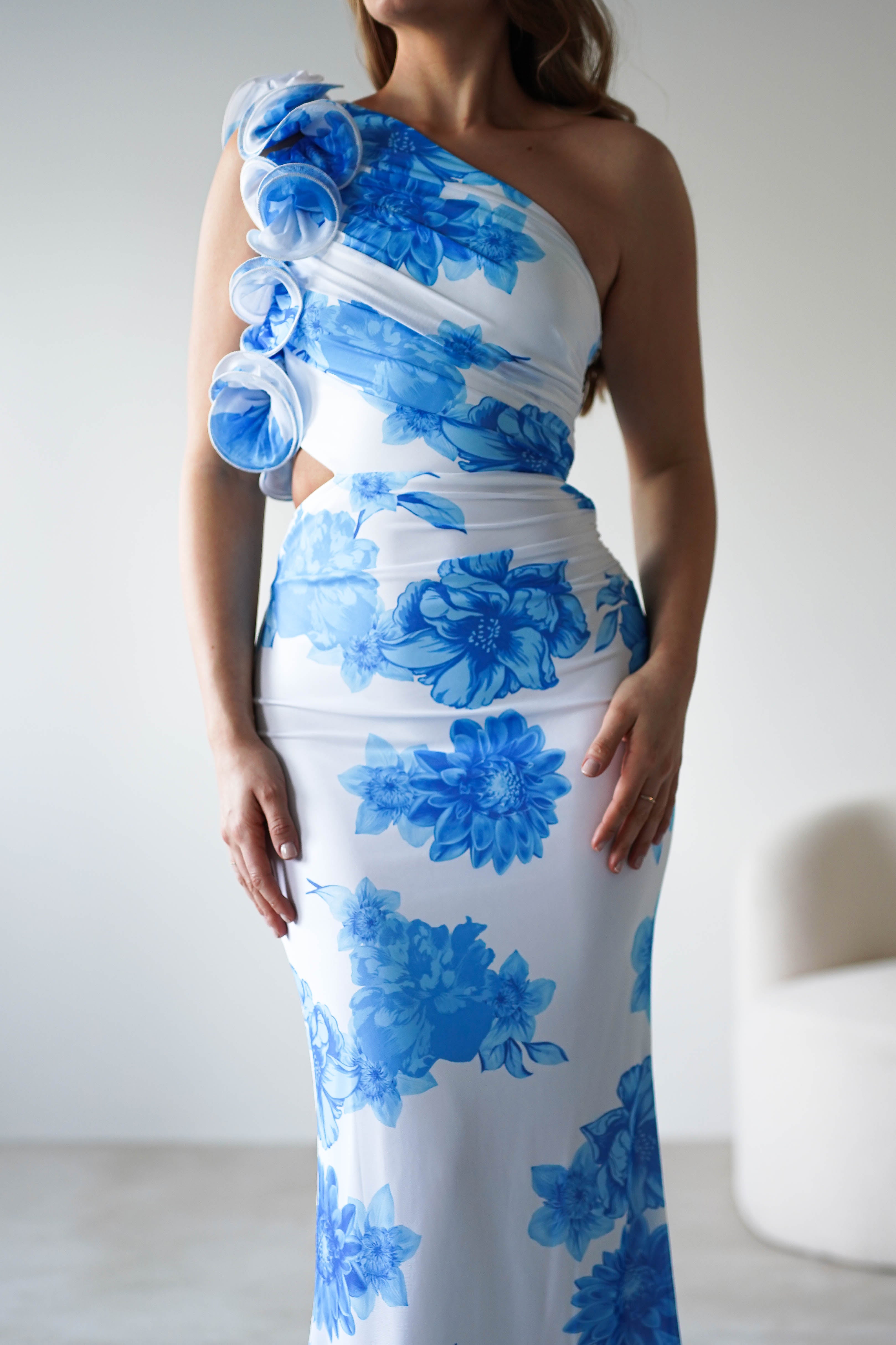 A woman in a white, one-shoulder wedding guest dress with bold summer blue print and delicate fabric flower details stands indoors. The fitted silhouette and elegant ruffle feature add a stylish touch to the sophisticated look.