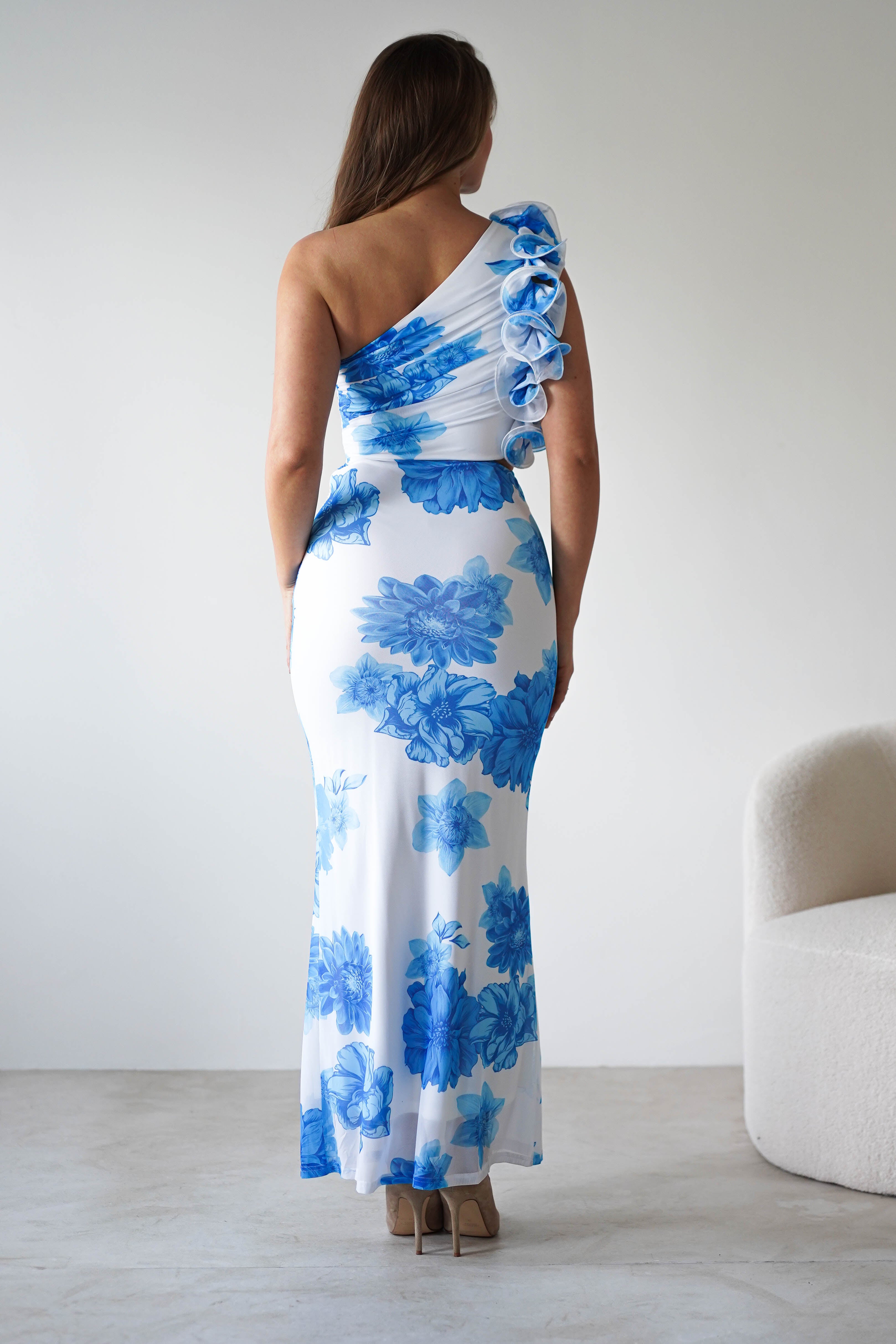 A woman stands facing away, wearing a summer blue print wedding guest dress with large ruffle feature on one shoulder and beige heels. She stands on a light floor beside a white chair.