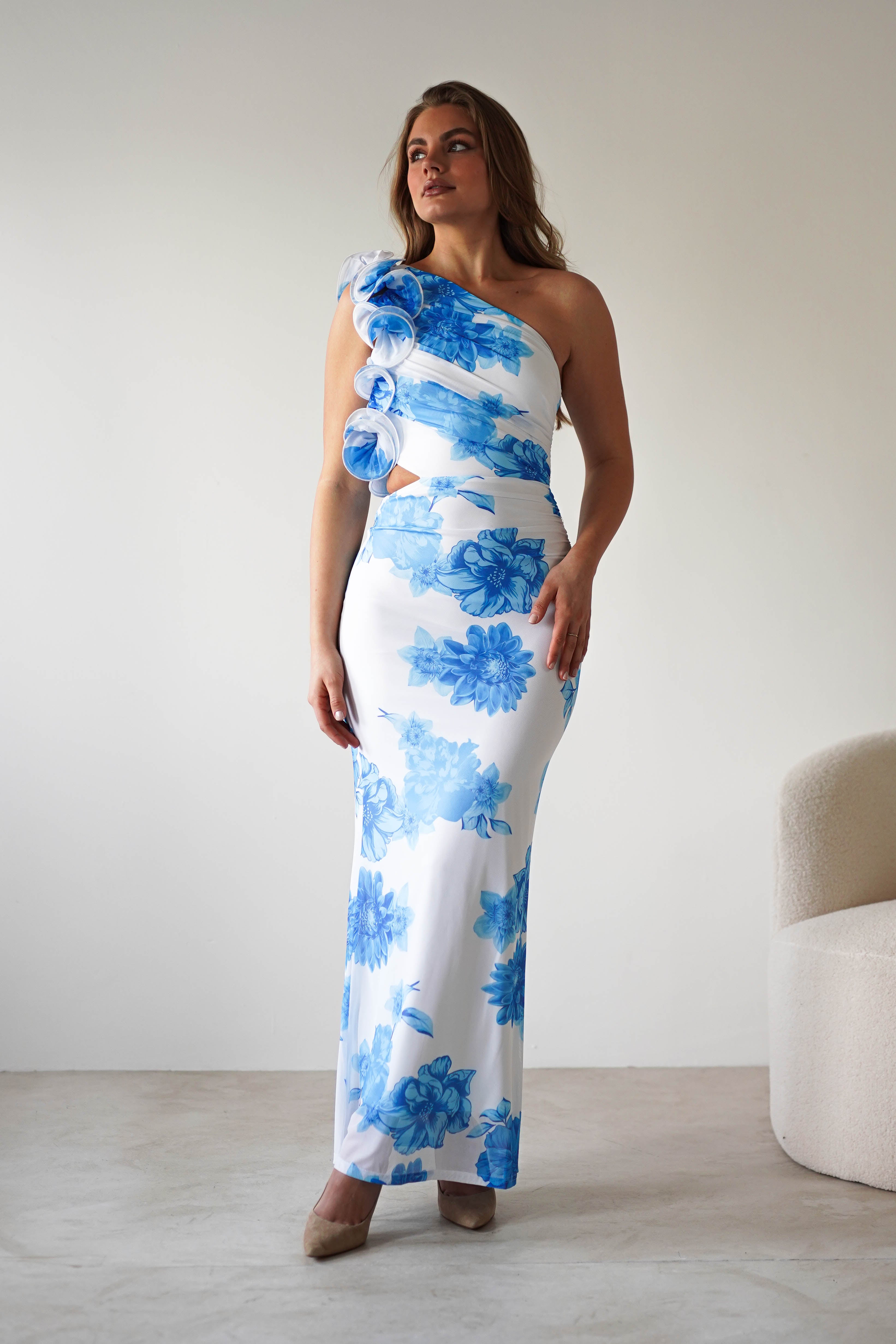 A woman wearing a fitted, one-shoulder wedding guest dress with a summer blue print and ruffle feature stands indoors next to a cream-colored chair, posing confidently.