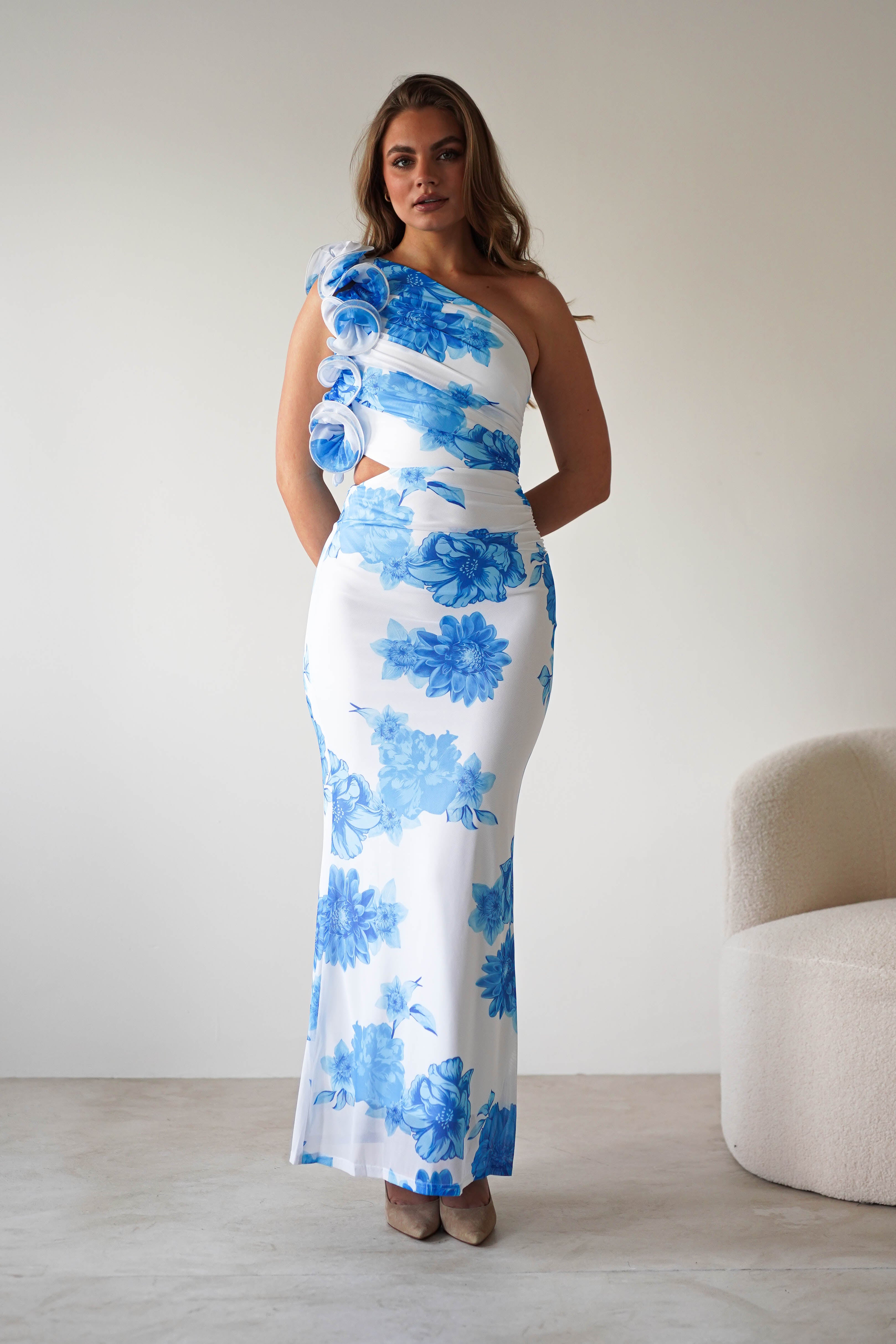 A woman stands indoors wearing a white, floor-length wedding guest dress with a summer blue print and a stylish one-shoulder ruffle feature. She has long hair and poses confidently next to a beige textured chair.