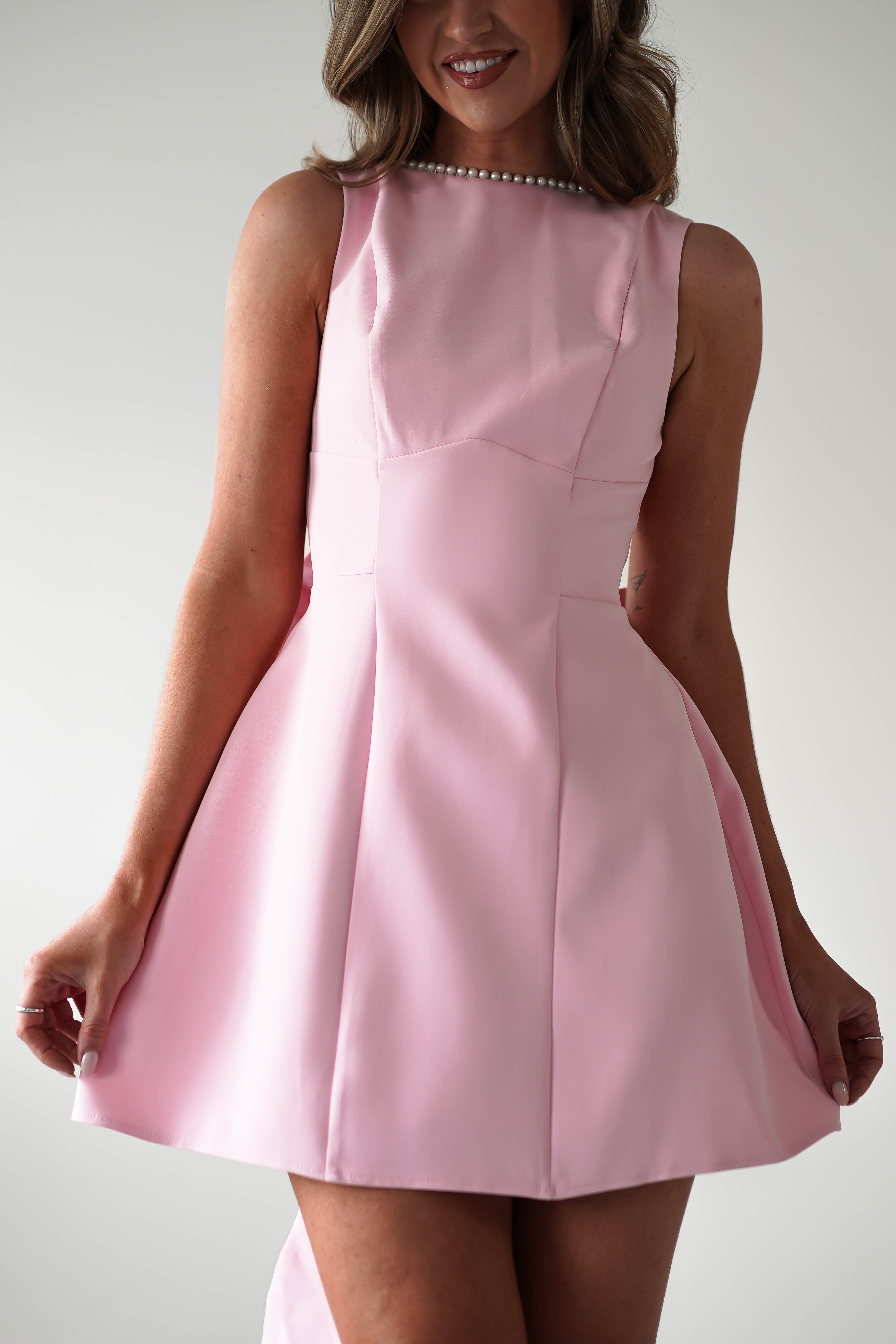 A woman wearing a sleeveless pink mini dress with a flared skirt and pearl trim along the neckline stands against a plain background, elegantly holding the sides of her dress—a perfect choice for a chic wedding guest dress.