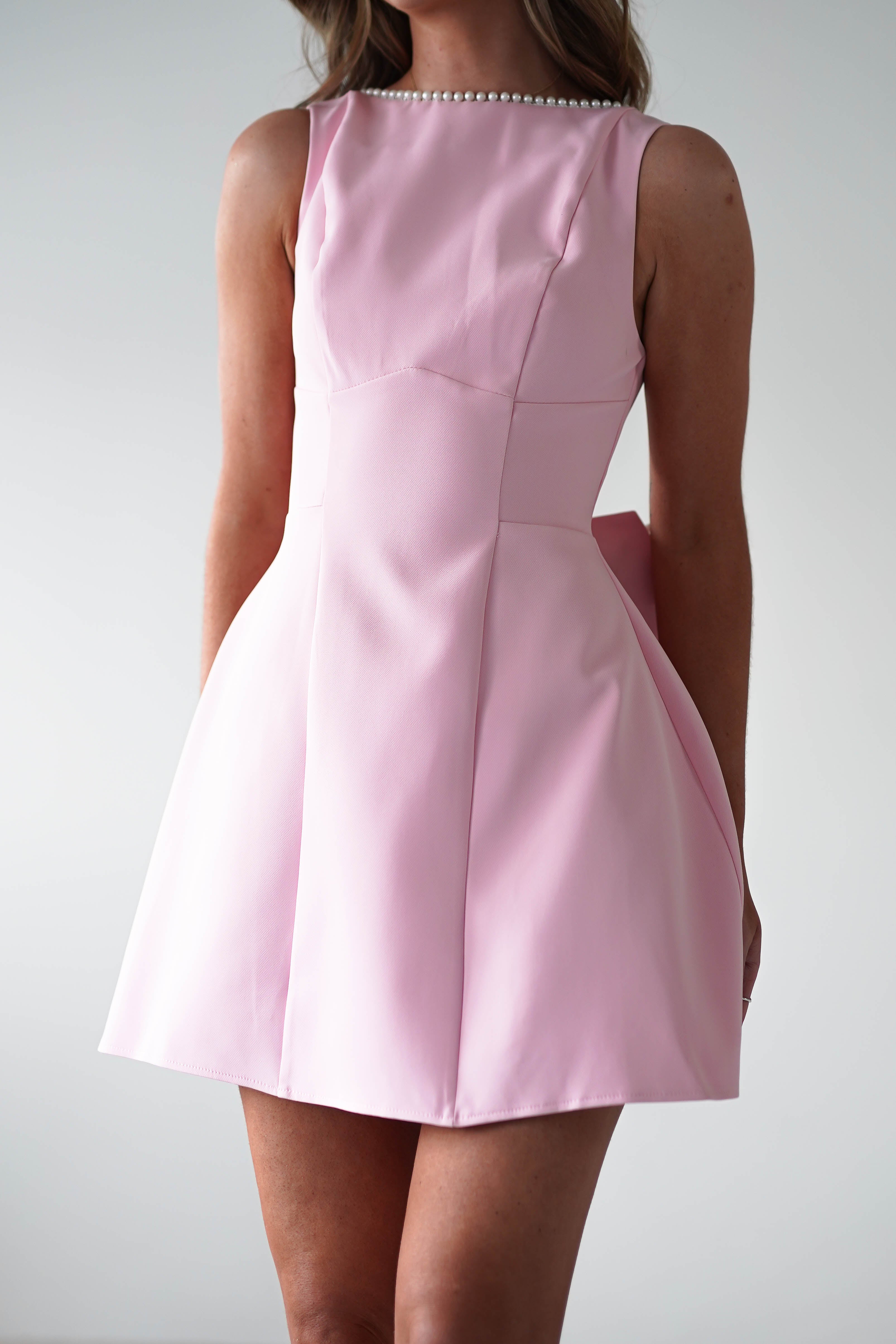 A woman stands against a plain light background in a sleeveless pink mini dress featuring pearl embellishments at the neckline. The flared skirt and fitted waist accentuate her style, making it a perfect choice for a wedding guest dress with its smooth texture and elegant design.