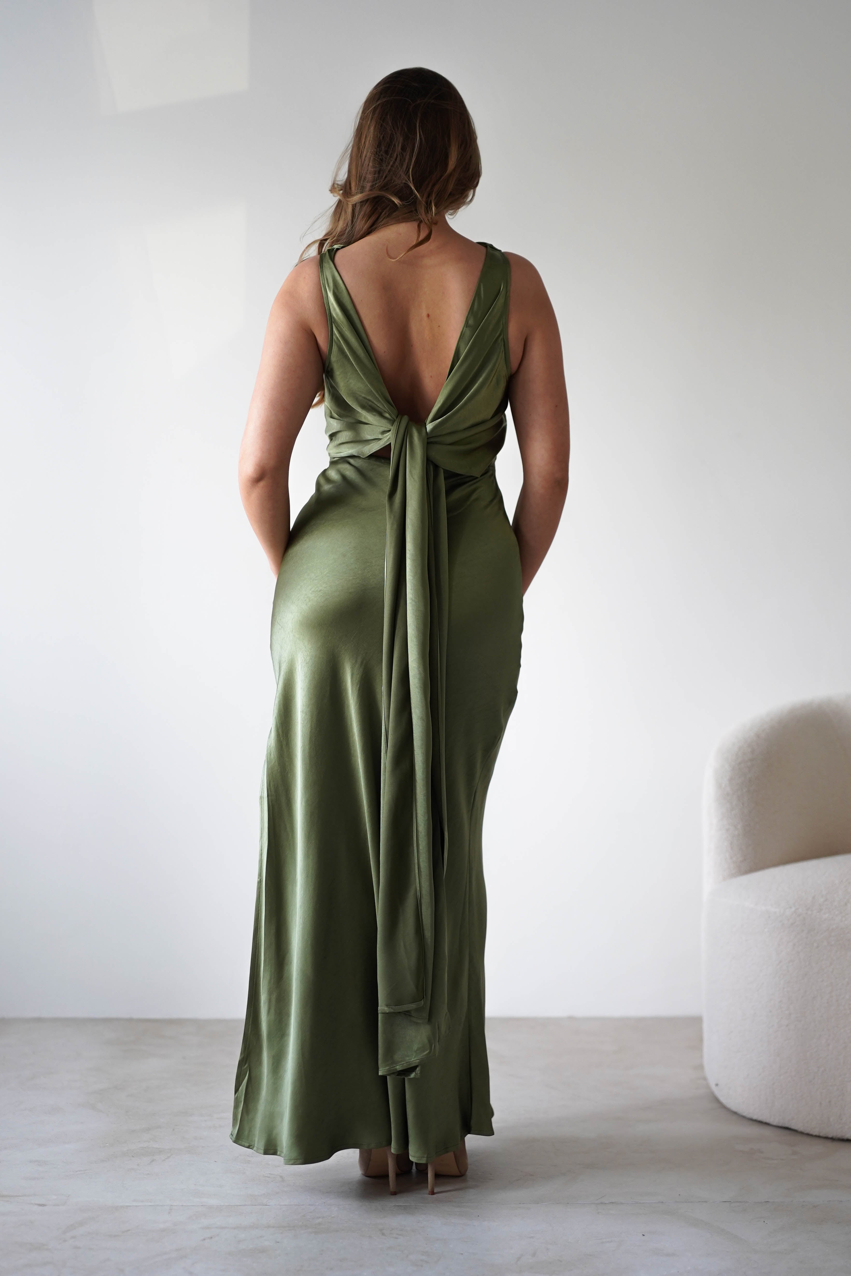 A woman stands facing away, wearing a Satin knit maxi dress in olive green with a low-cut open back and bow detail. Perfect for a wedding guest, she stands on a light floor near a modern white armchair against a plain background.