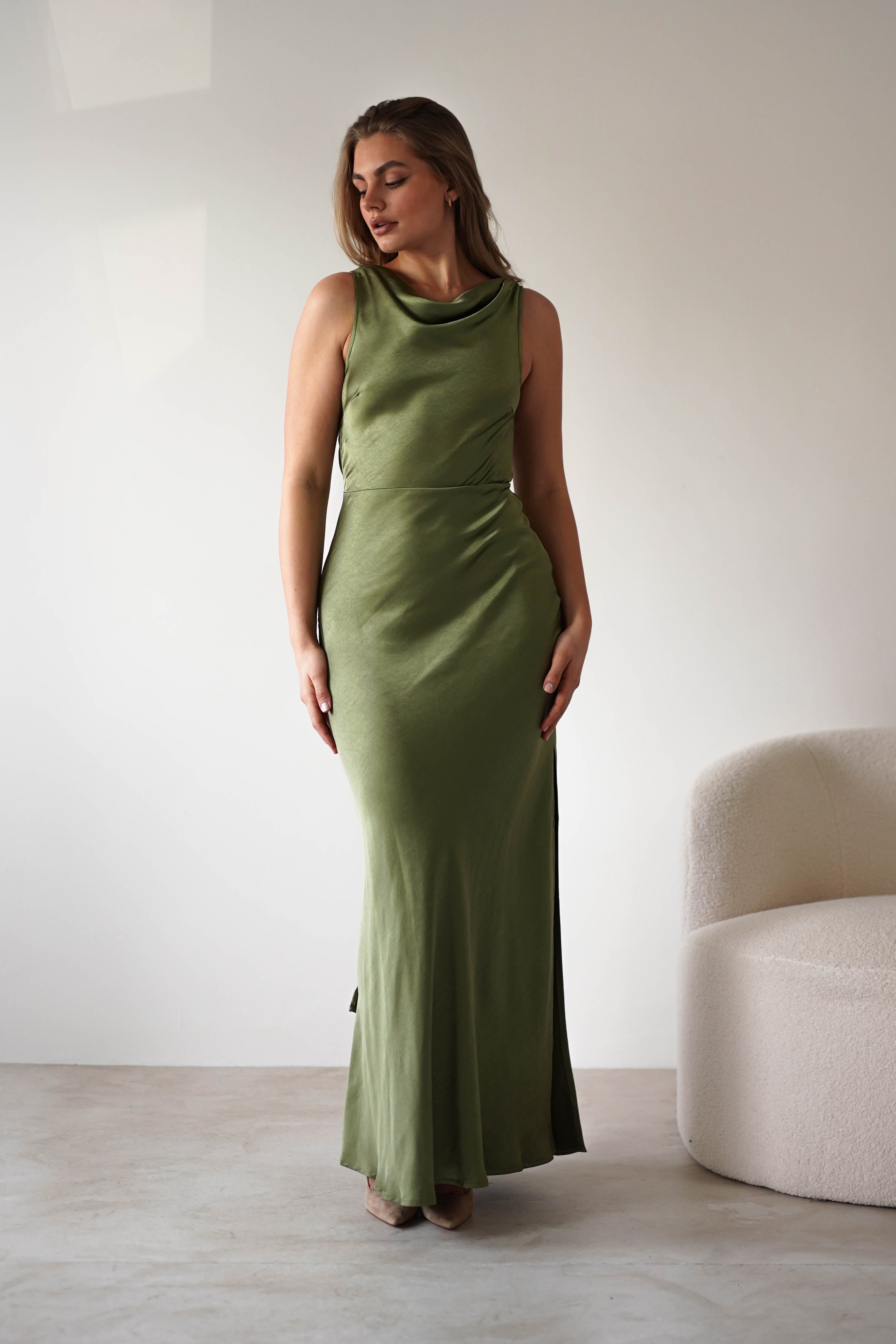A woman stands indoors in a long, sleeveless olive green satin knit maxi dress with a low-cut open back. She poses next to a cream-colored modern chair, her hair down, looking slightly to the side against a plain light background.