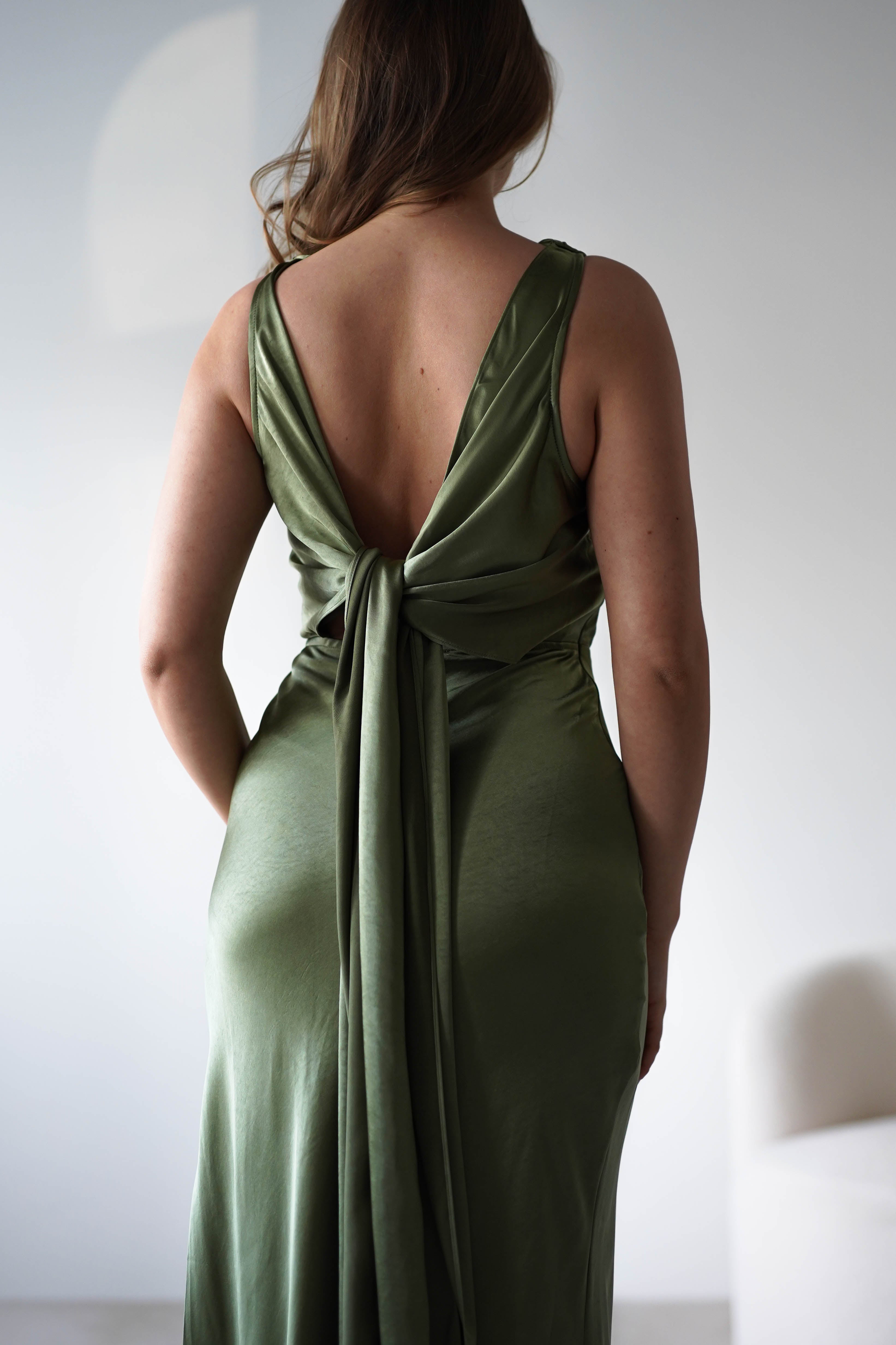Woman in a silky, olive green Satin knit maxi dress with a low-cut open back and long ties stands facing away in a softly lit, minimalist room with white walls and a light-colored chair partially visible—perfect for a chic wedding guest look.