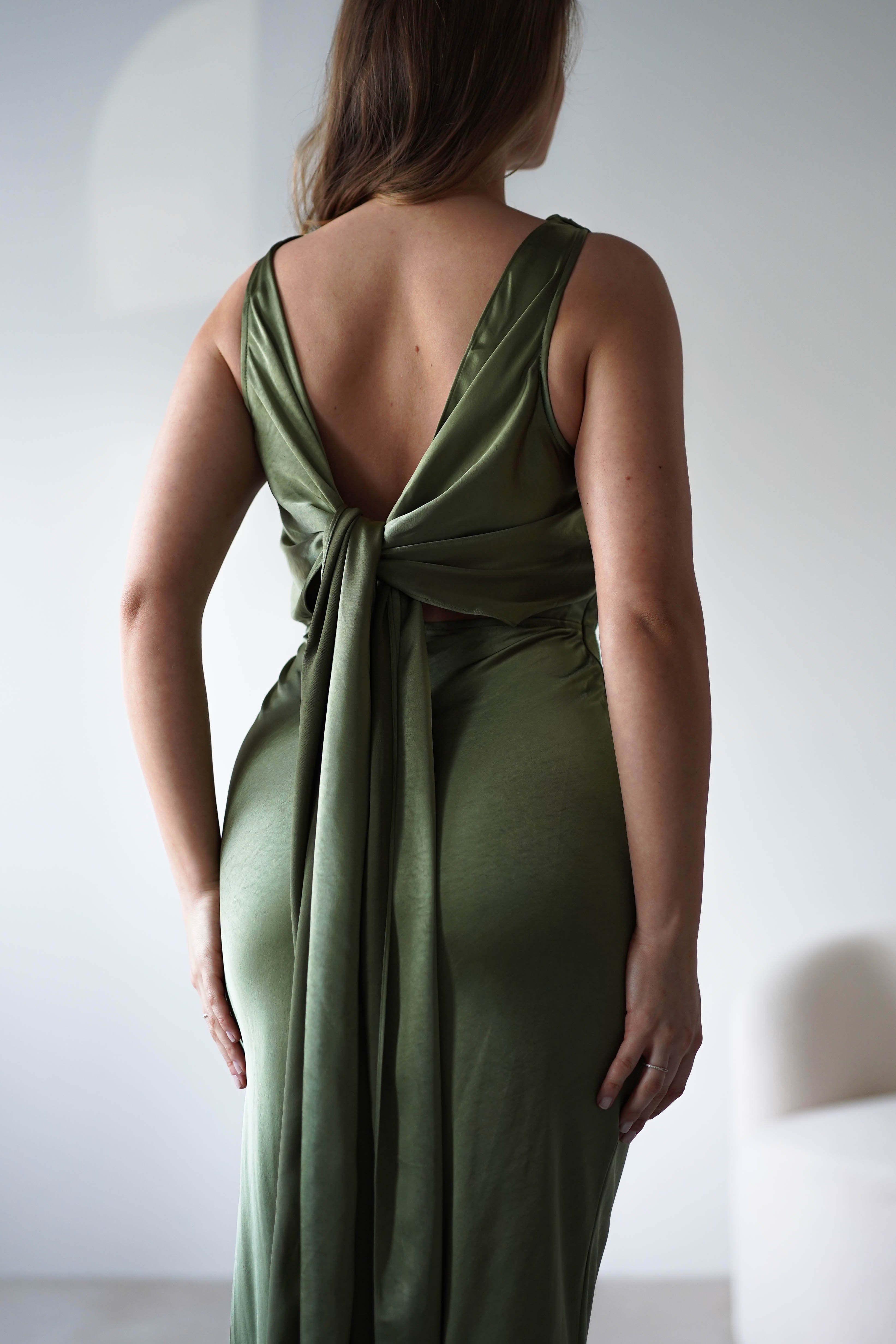 A woman wearing a sleeveless, olive green satin knit maxi dress with a low-cut open back and long, draped ties stands facing away in a softly lit room with a minimal background—perfect for a wedding guest look.