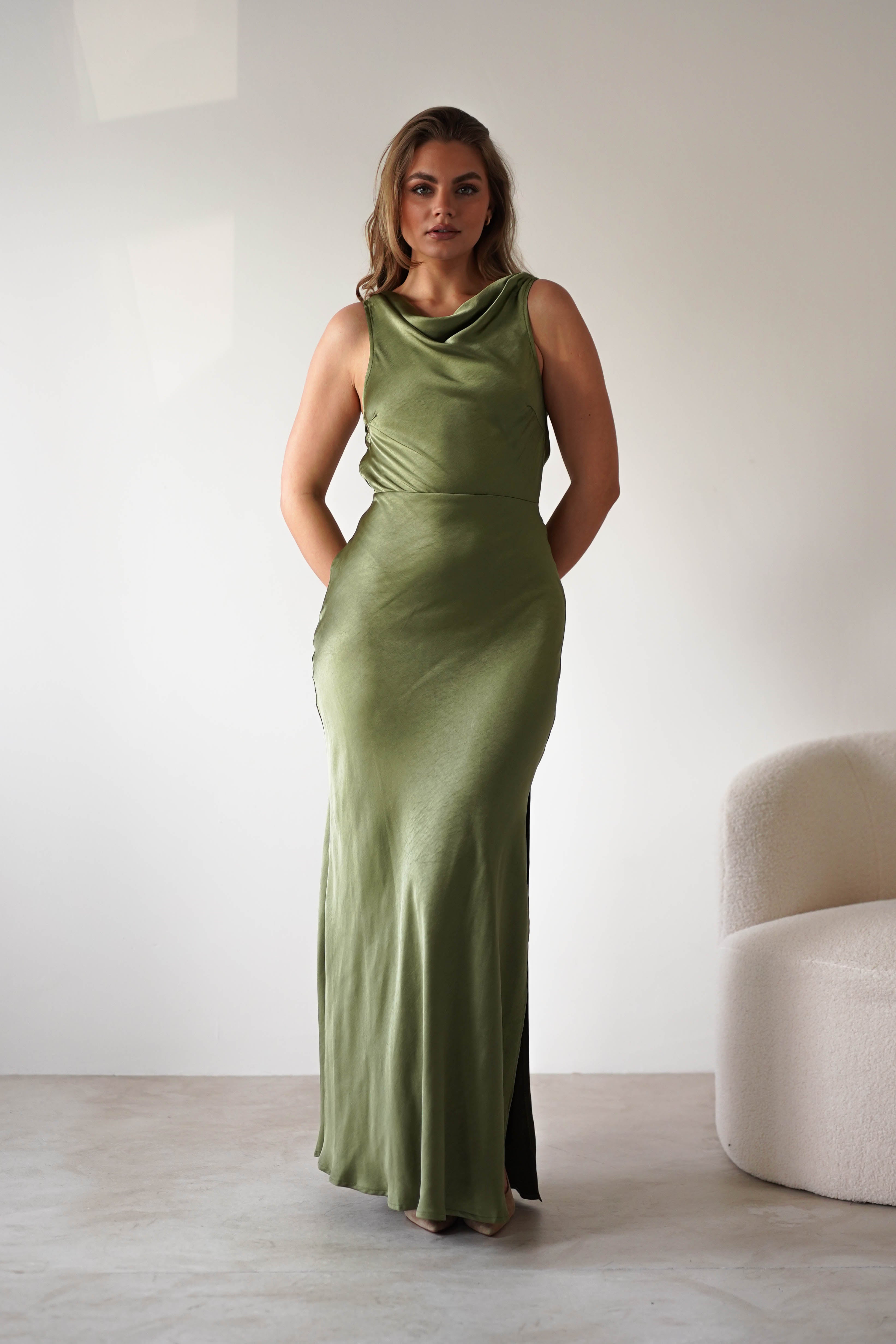 A woman stands indoors wearing a sleeveless, olive green satin knit maxi gown with a draped neckline and low-cut open back, her hands in the dress pockets. She is next to a cream-colored armchair against a plain white wall—perfect for a wedding guest look.