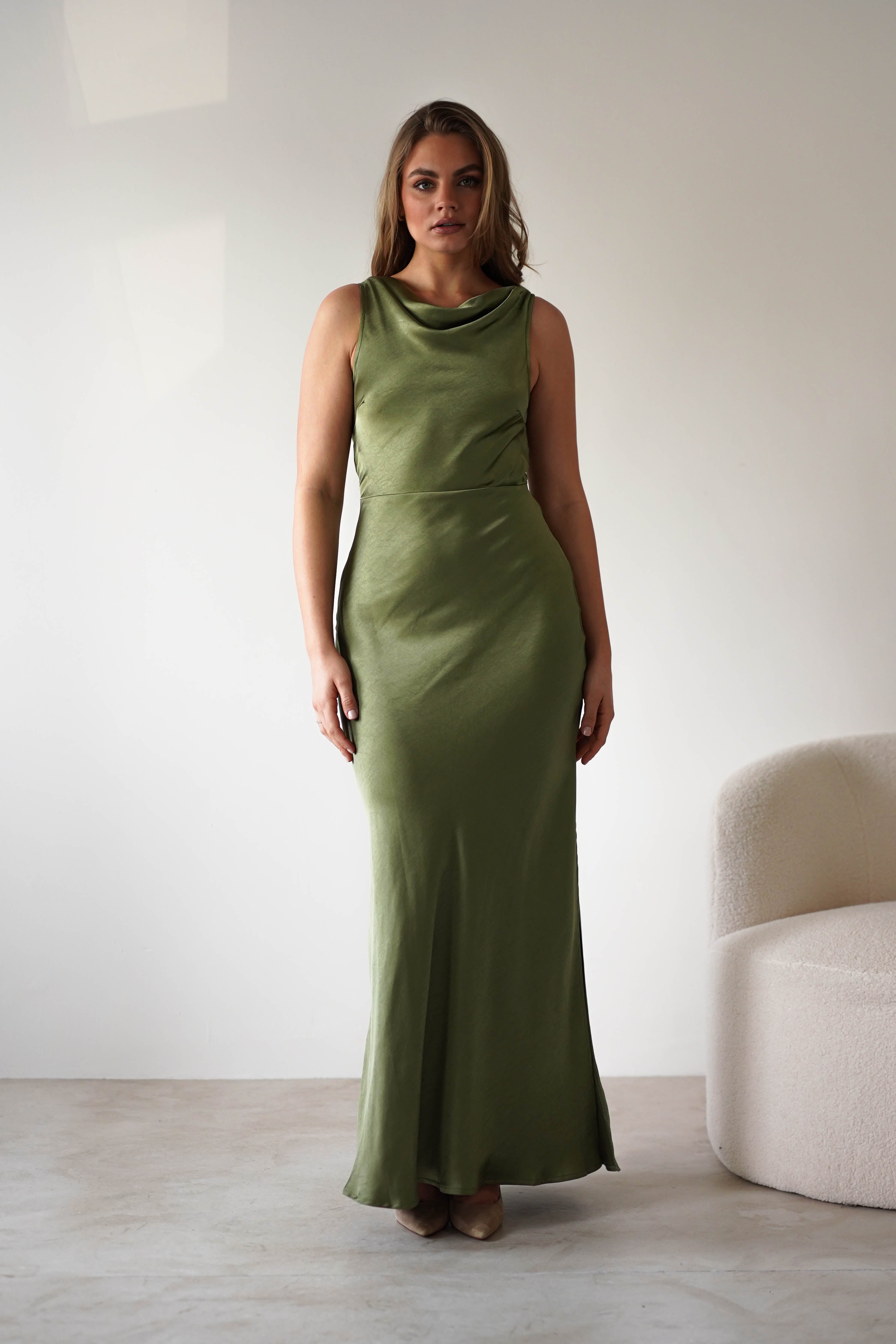 A woman stands indoors in a sleeveless satin knit maxi dress with a low-cut open back, perfect for a wedding guest. She has light brown hair and poses beside a white, curved armchair against a plain, light-colored wall.