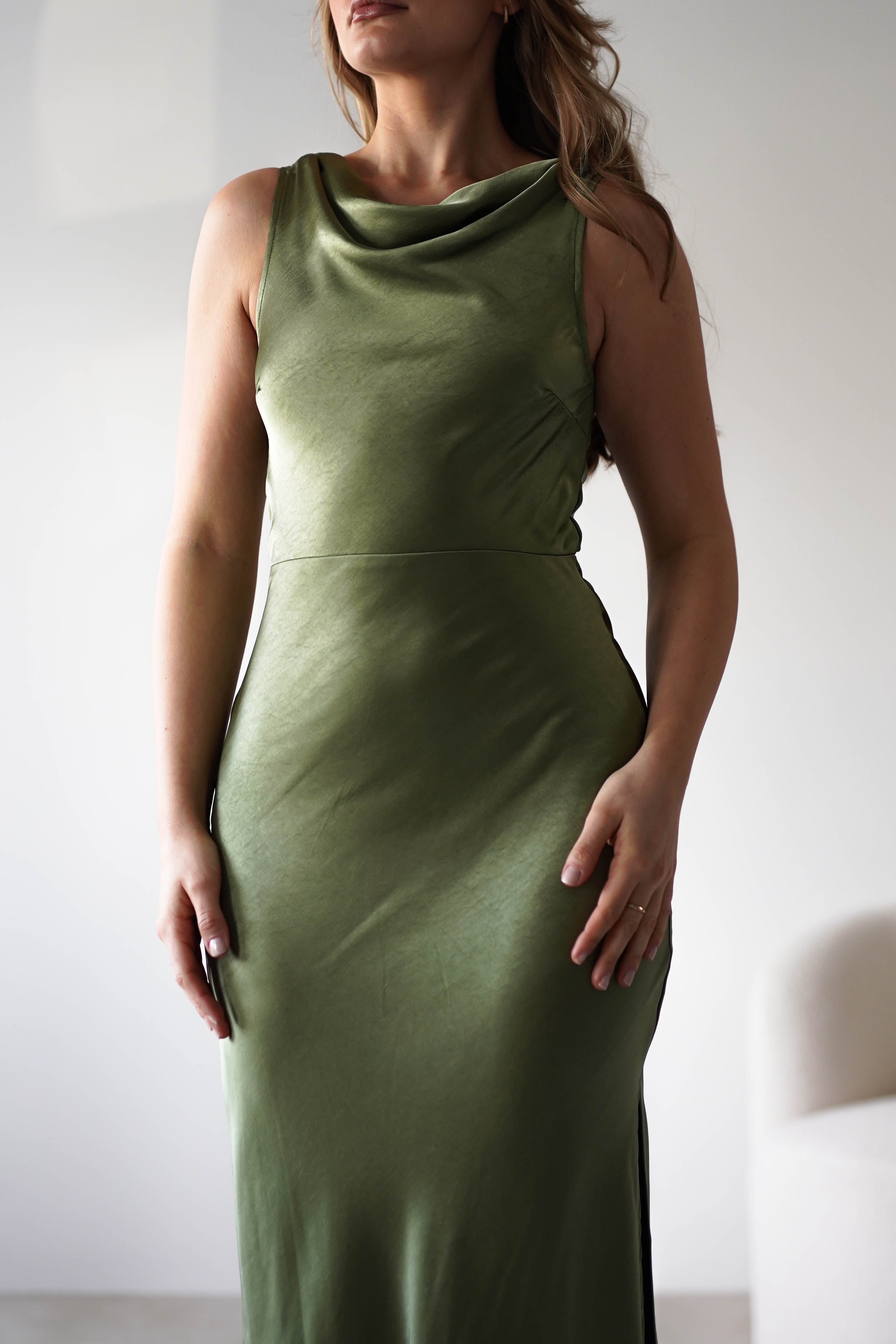 A woman wearing a sleeveless, olive green satin knit maxi dress with a low-cut open back stands indoors against a plain light background, her hands resting gently by her sides—perfect for a stylish wedding guest.