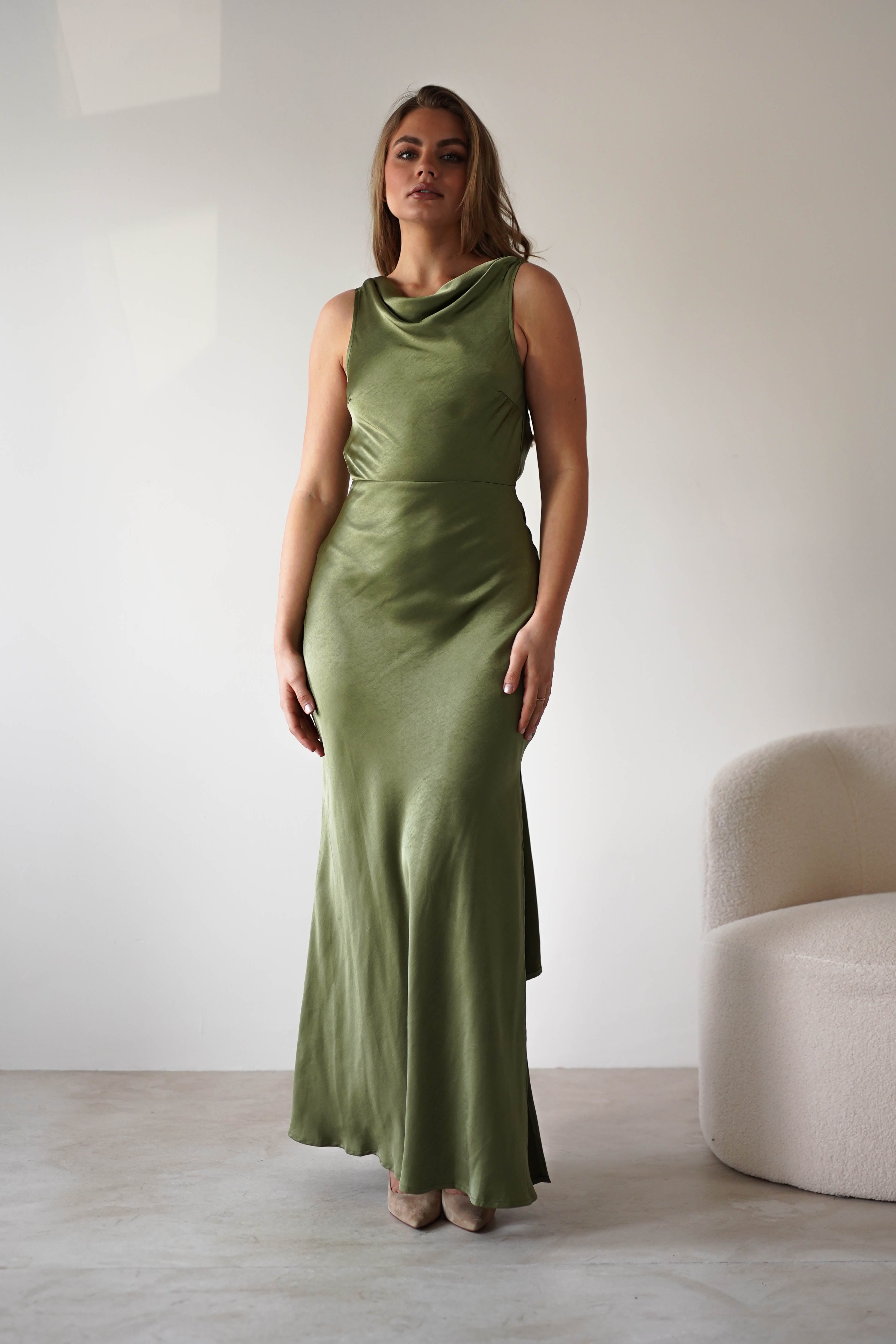 A woman wearing a sleeveless, olive green satin knit maxi gown with a low-cut open back stands indoors near a white curved chair, against a plain light-colored wall—perfect for a stylish wedding guest look. She has long hair and a neutral expression.