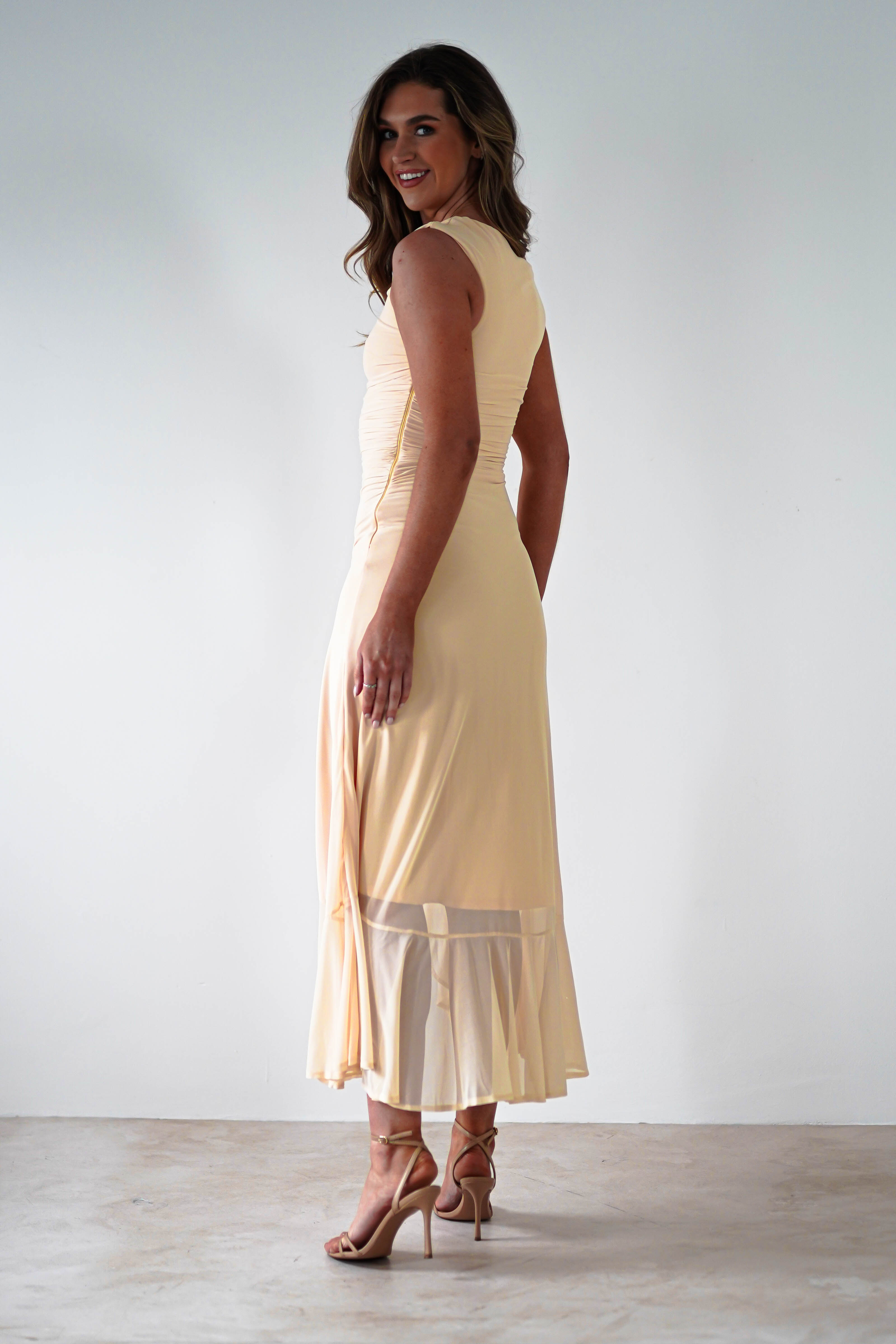 A woman poses in a sleeveless, light beige midaxi dress with stretchy mesh fabric detailing at the hem. Shes wearing nude high-heeled sandals and stands against a plain white background, looking over her shoulder, perfect for special occasions.