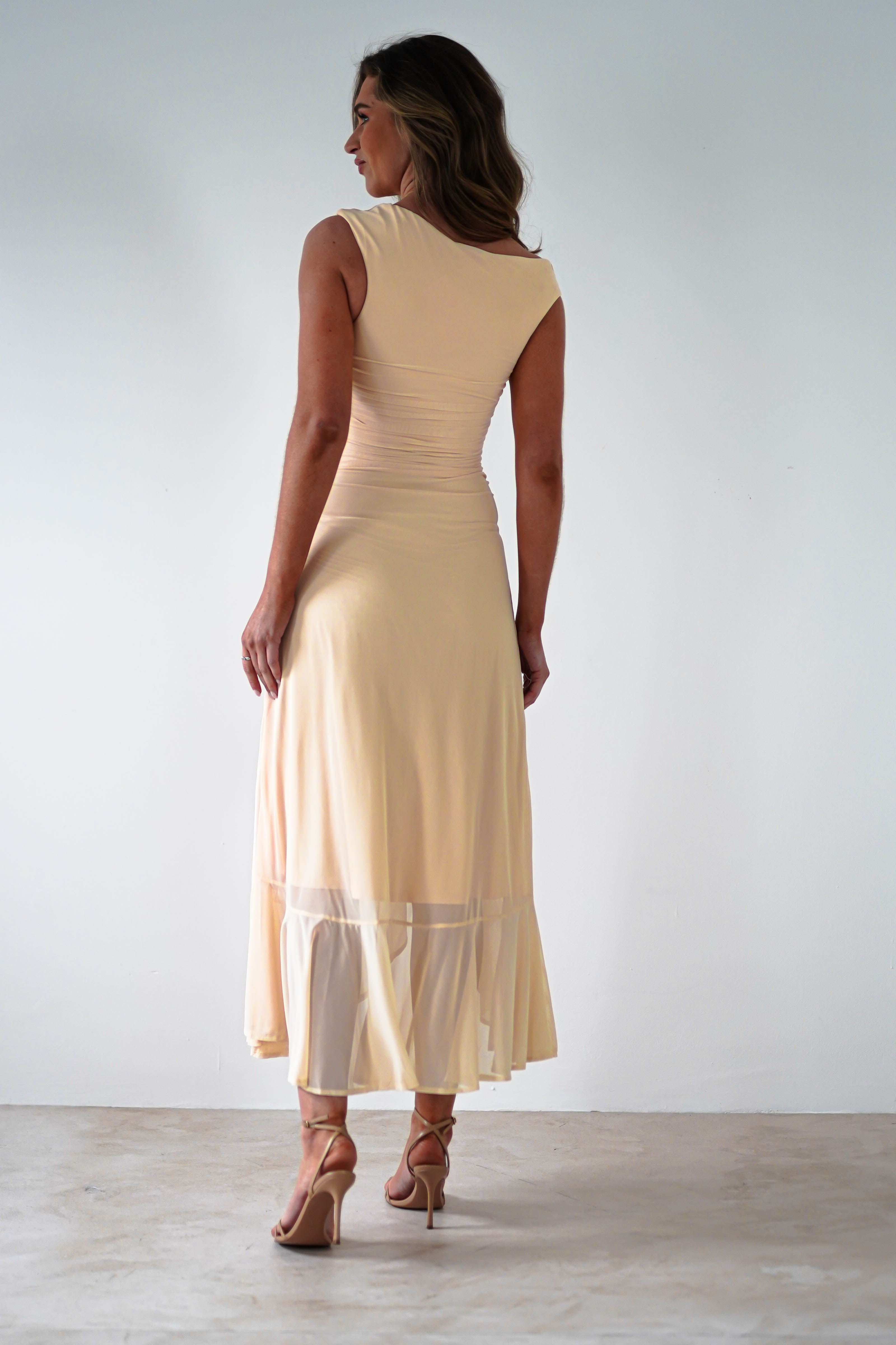 A woman in a light beige, stretchy mesh midaxi dress stands with her back turned. The dress boasts a fitted top and flowing skirt with a sheer hem, perfect for special occasions. She wears high-heeled sandals and stands on a light-colored floor against a plain wall.