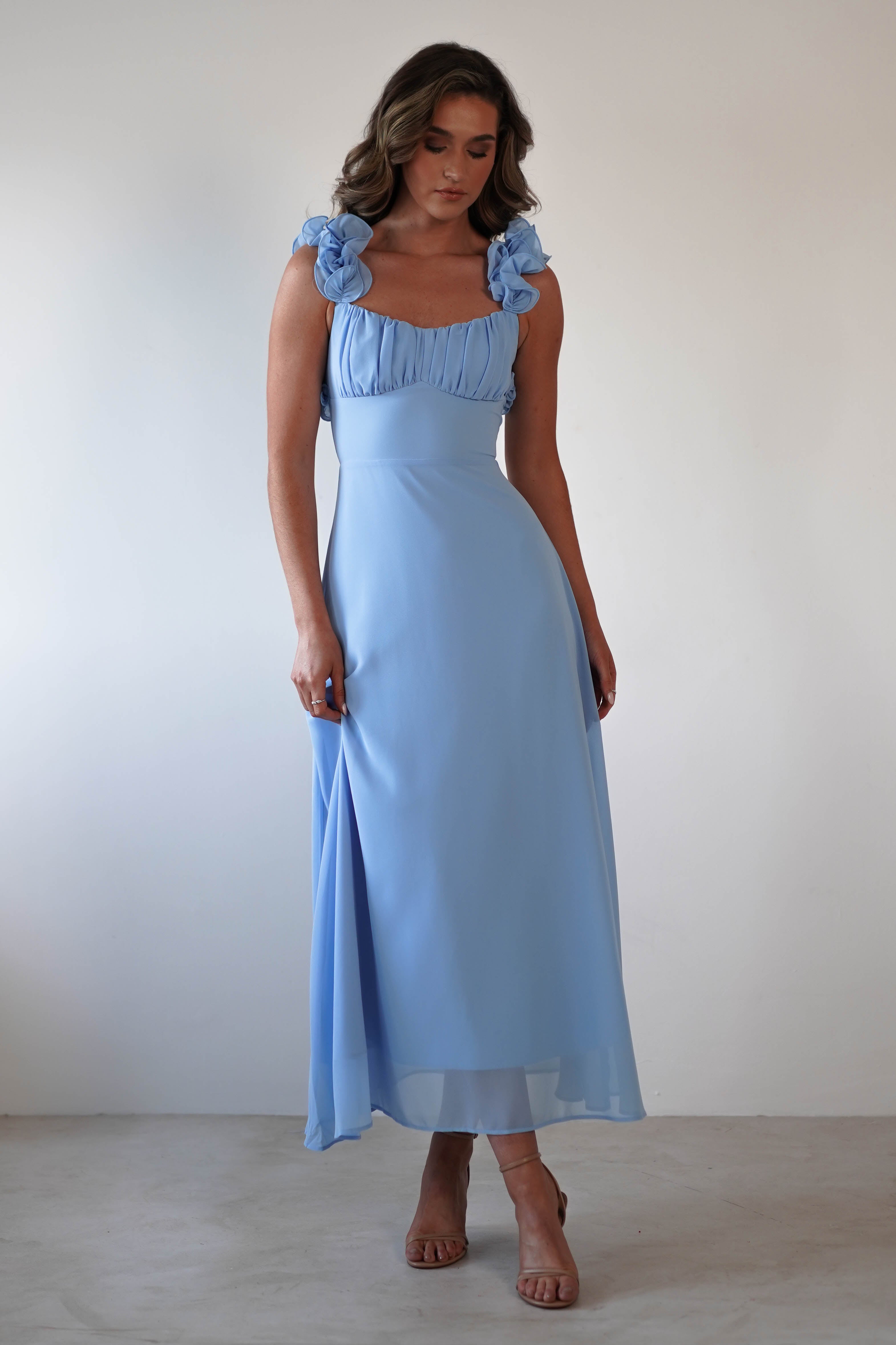 A woman in a light blue chiffon midaxi dress with floral ruffle details on the straps stands against a plain white background. She has long, wavy hair and is wearing strappy heels, perfectly embodying the elegance of a wedding guest.