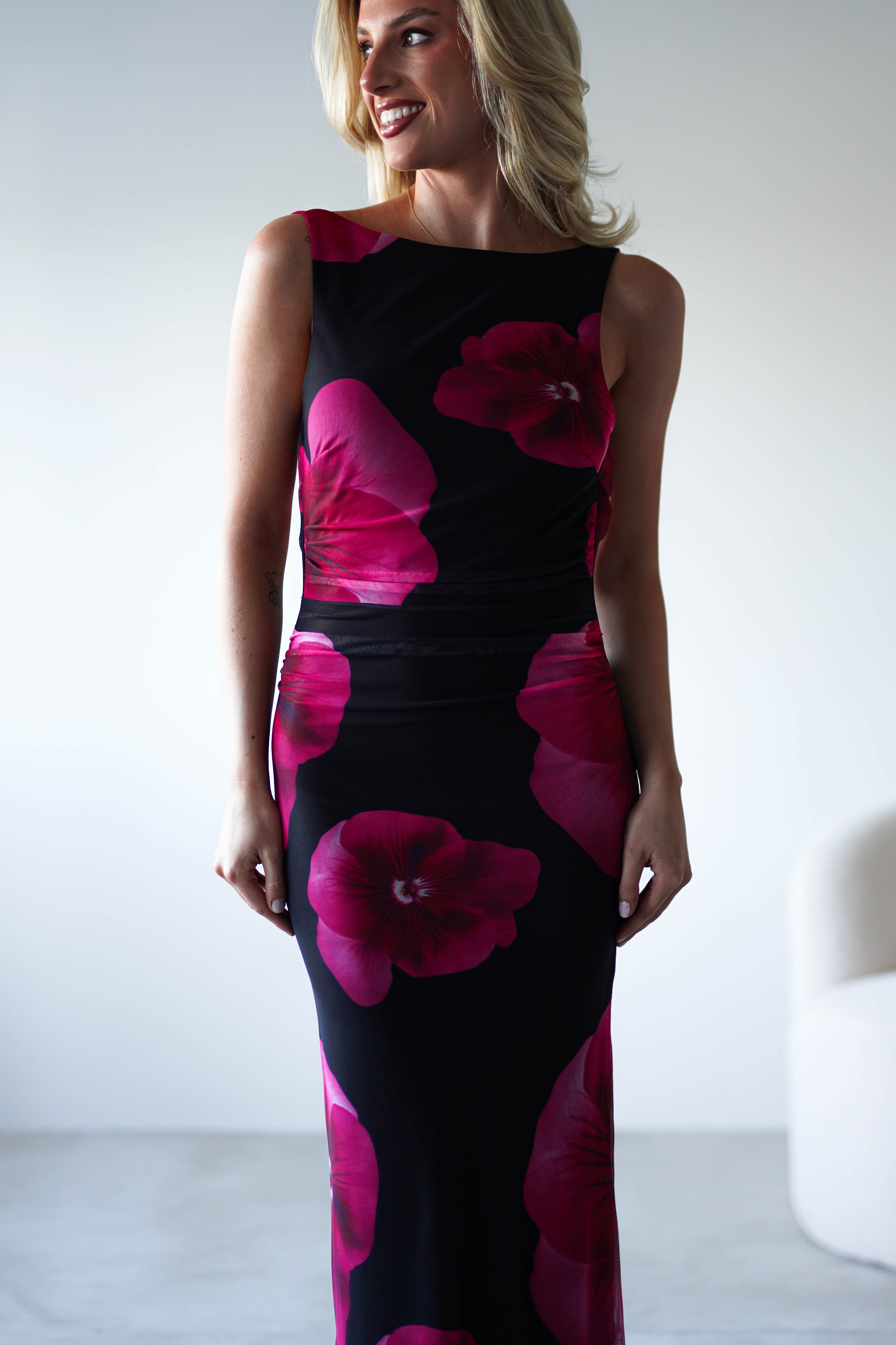 A woman stands indoors in a sleeveless black and red floral print maxi dress, smiling and looking to the side. The softly lit, minimal background features a partially visible white chair—perfect attire for a wedding guest.