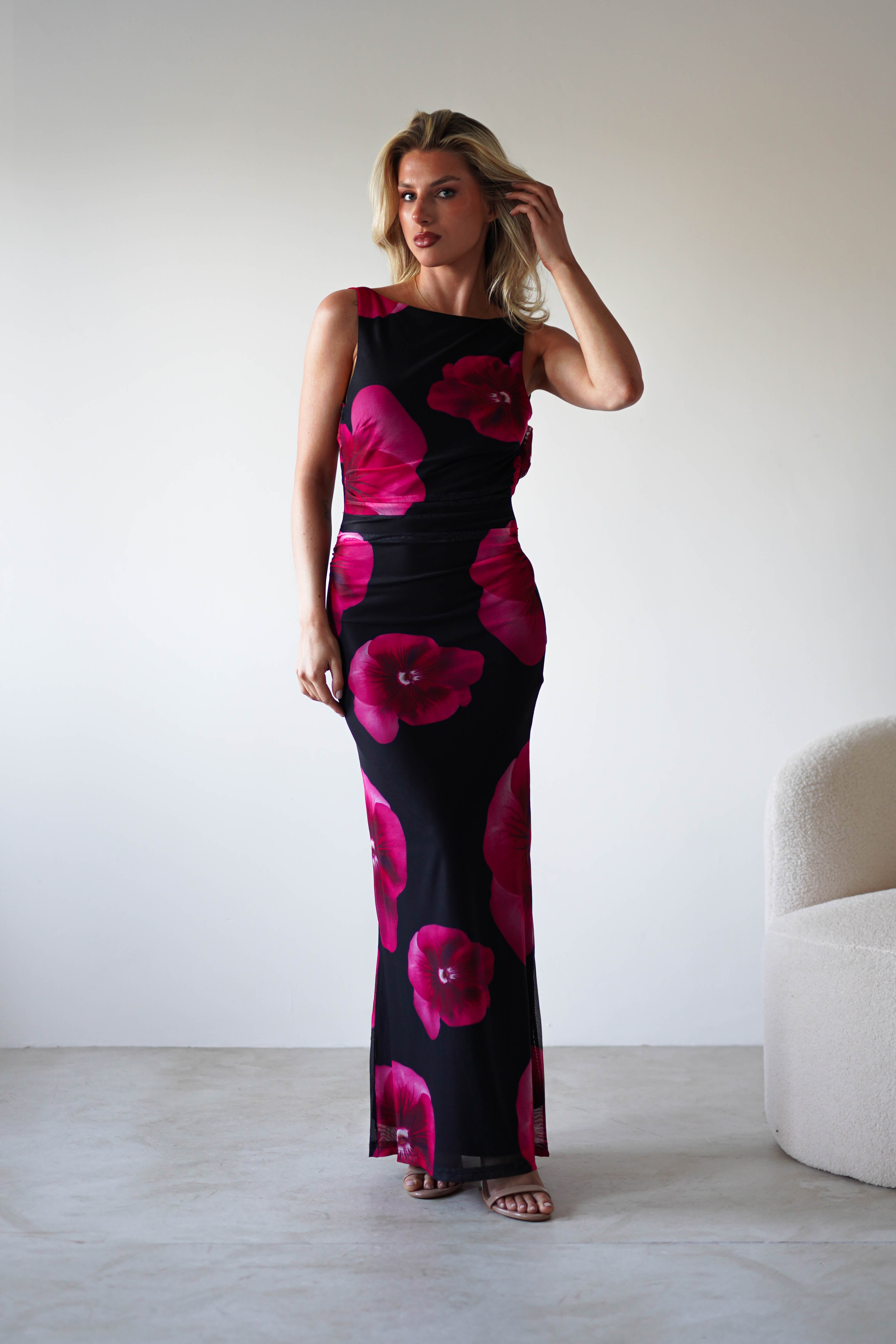 A woman stands indoors against a white wall, wearing a sleeveless black and red floral print maxi dress and beige heeled sandals—an elegant look perfect for a wedding guest. She touches her hair with one hand and gazes slightly to the side.
