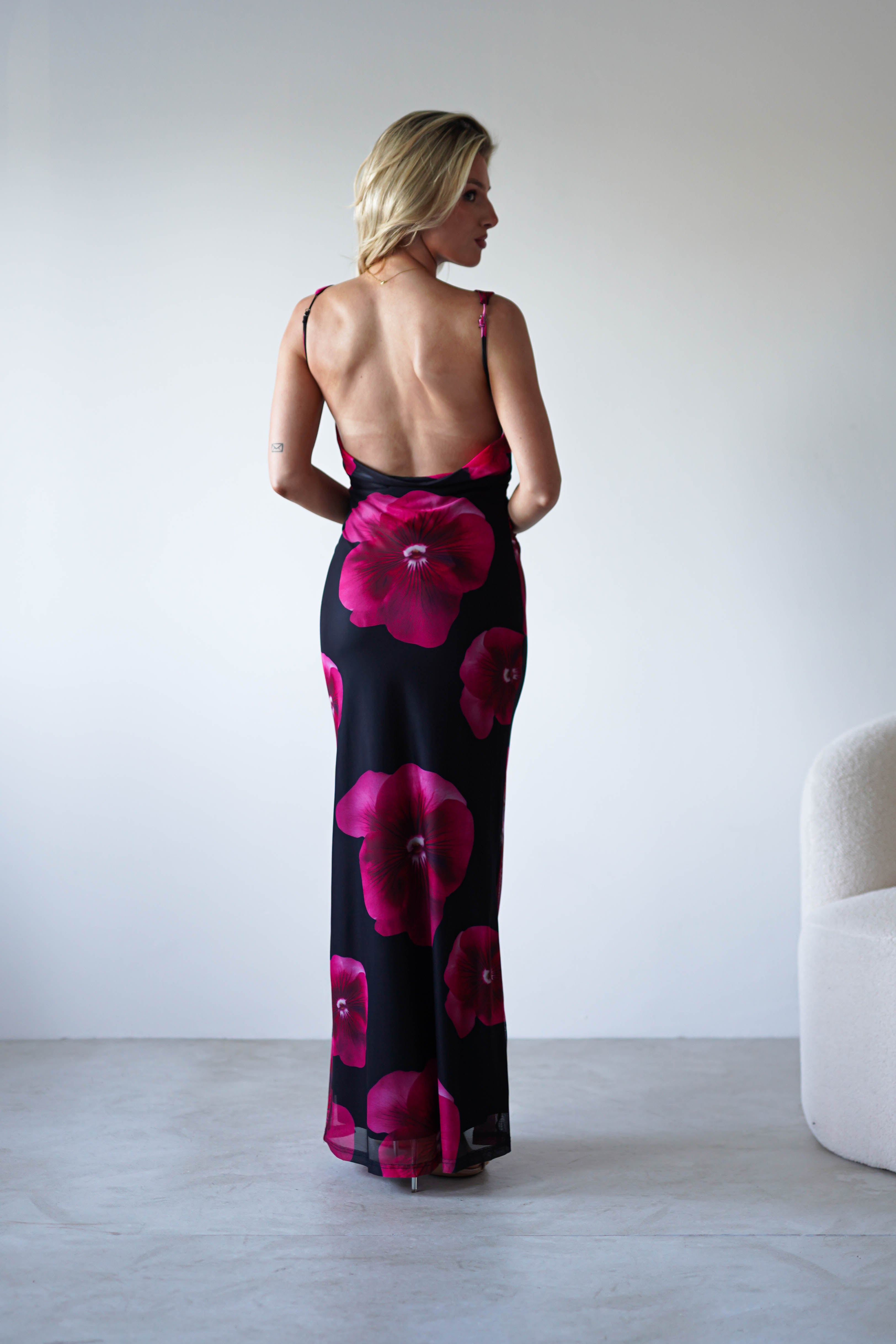 A woman with blonde hair stands facing away, wearing a black and red floral print maxi dress with an open back—perfect for a wedding guest. She is indoors in a minimalist room with light walls and a white textured chair nearby.