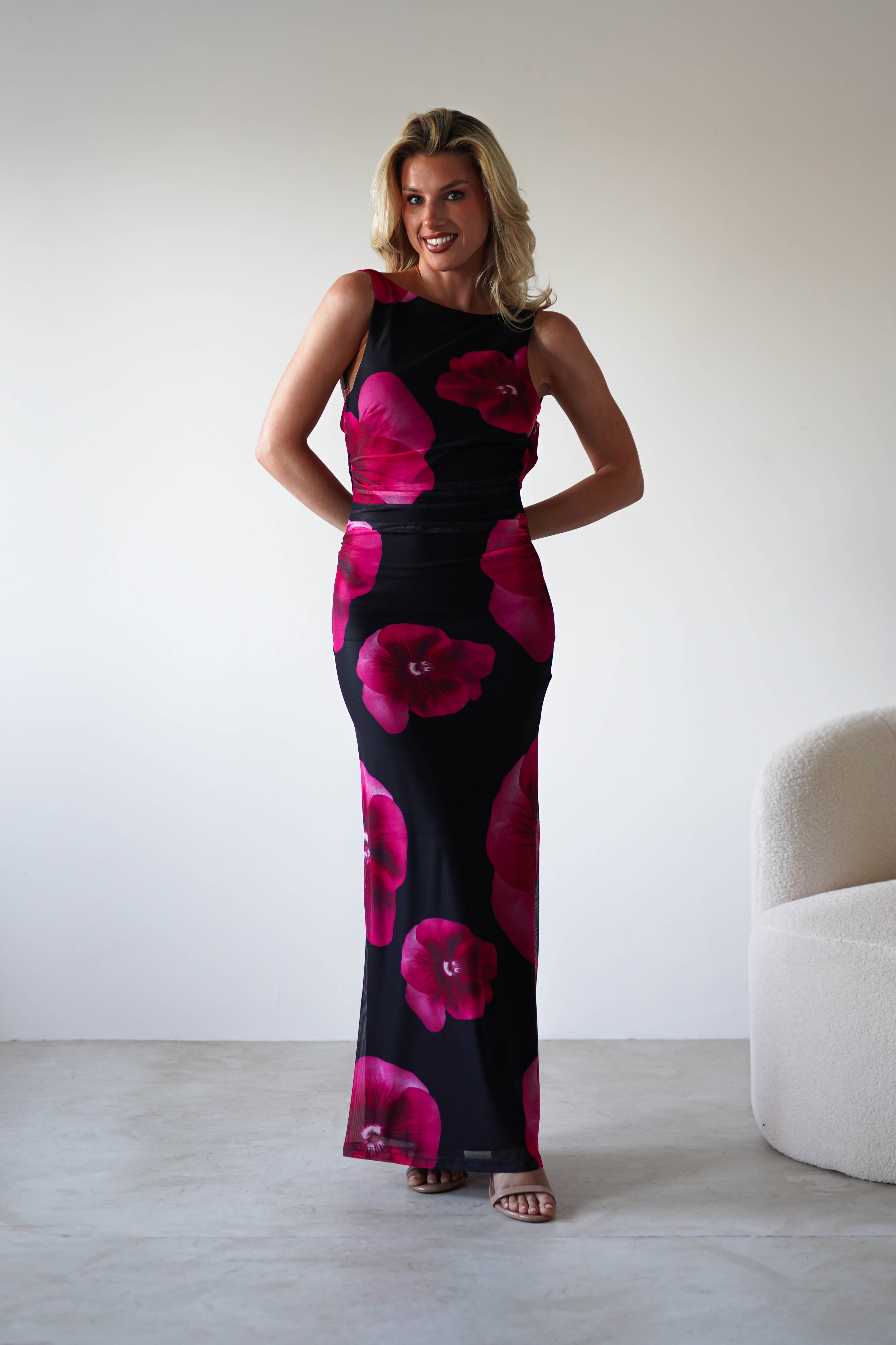 A woman stands indoors wearing a sleeveless black and red floral print maxi dress, smiling with her hands behind her back. She is next to a light-colored curved chair against a plain white wall—perfect attire for a stylish wedding guest.