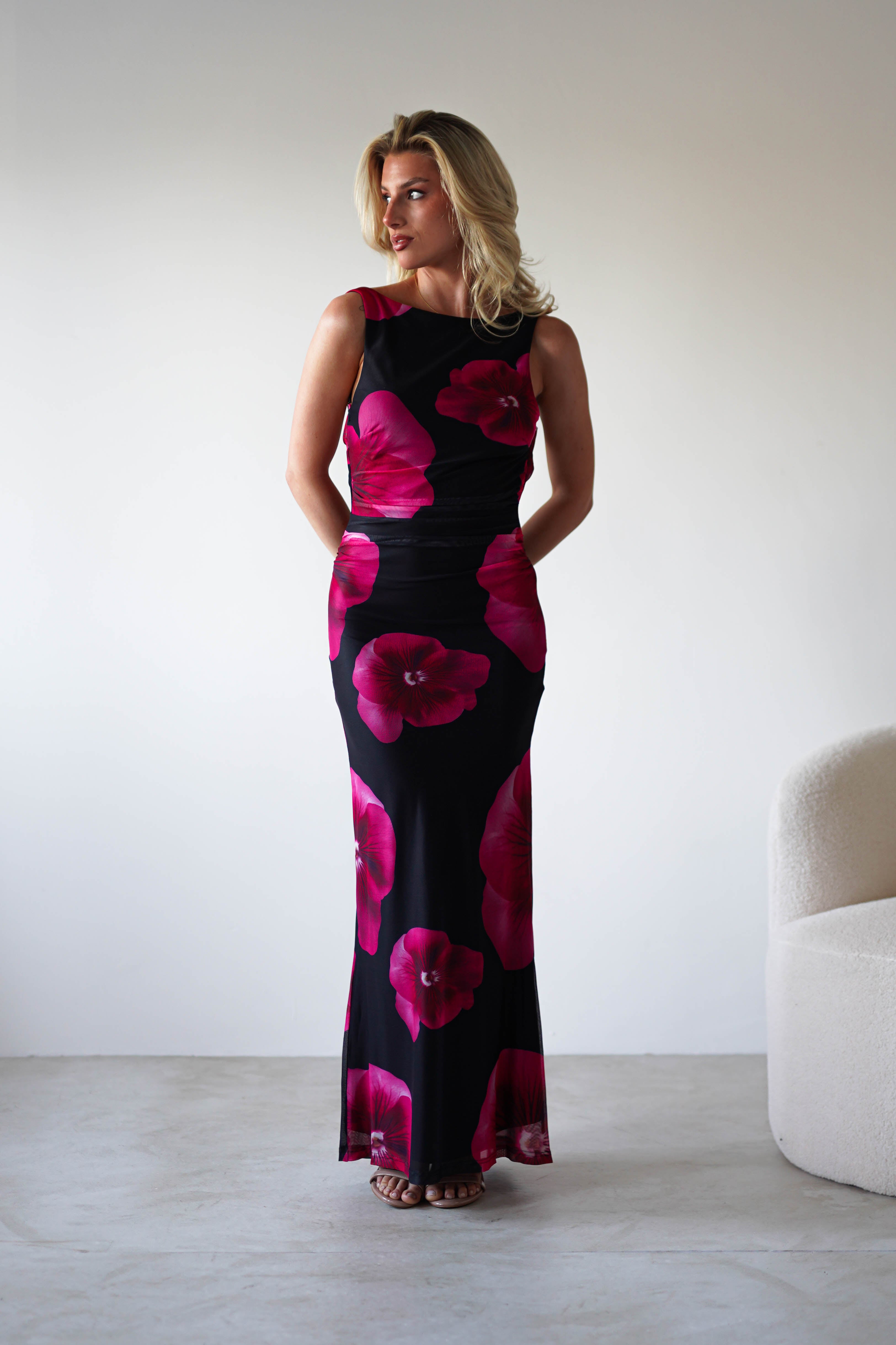 A woman with blonde hair stands indoors in a black and red floral print maxi dress, her hands behind her back, next to a white curved chair—an elegant look perfect for a wedding guest.