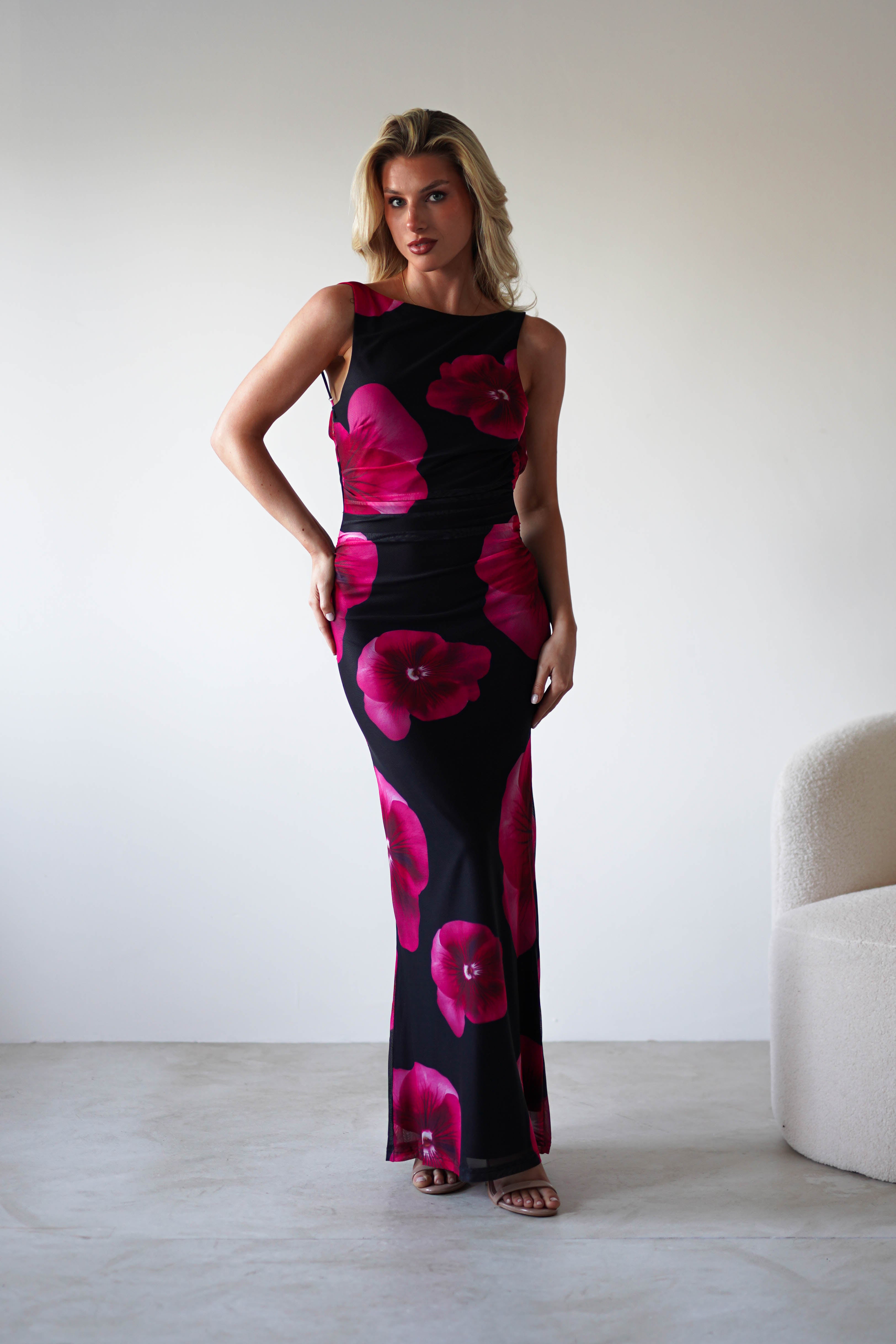 A woman stands indoors in a sleeveless, form-fitting black and red floral print maxi dress. With shoulder-length blonde hair, she poses near a light-colored chair against a plain white wall—an elegant look for a wedding guest.