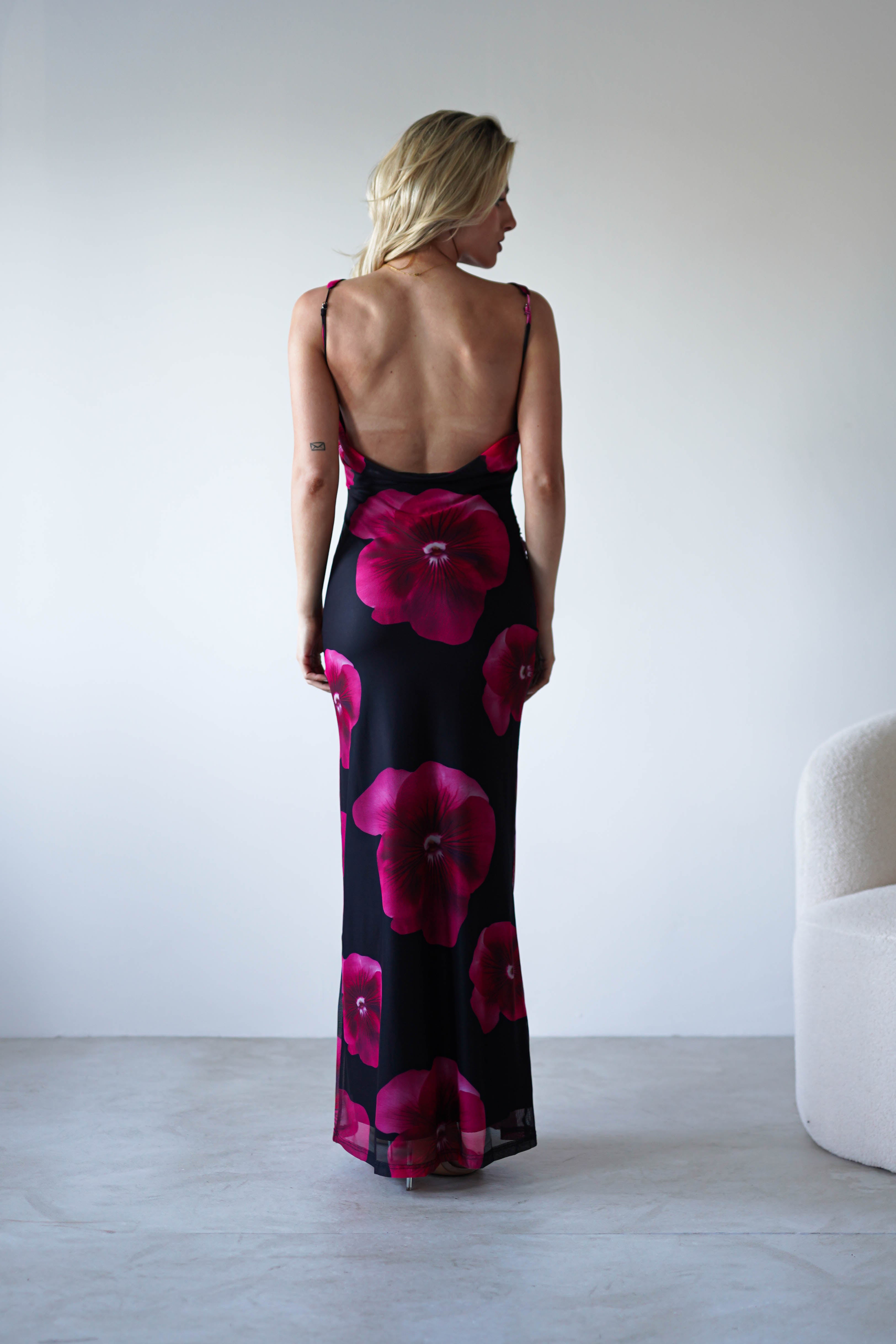 A woman with blonde hair stands facing away in a minimalistic room, wearing a black and red floral print maxi dress with an open back—an elegant choice for a wedding guest.