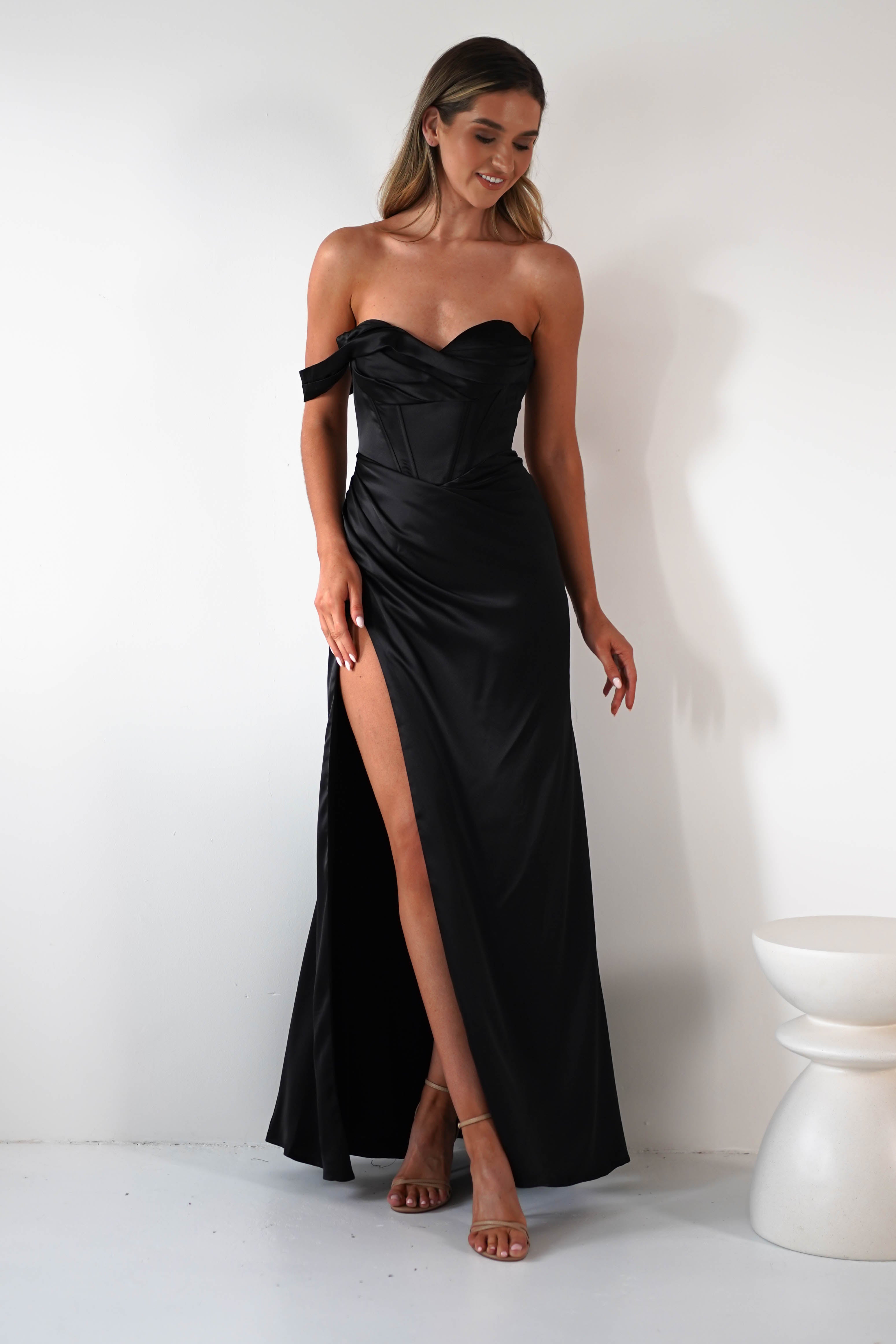 A woman in a strapless black satin corset dress with a high side slit poses against a white wall. She has long hair and wears high-heeled sandals, with one hand holding the side of her dress.