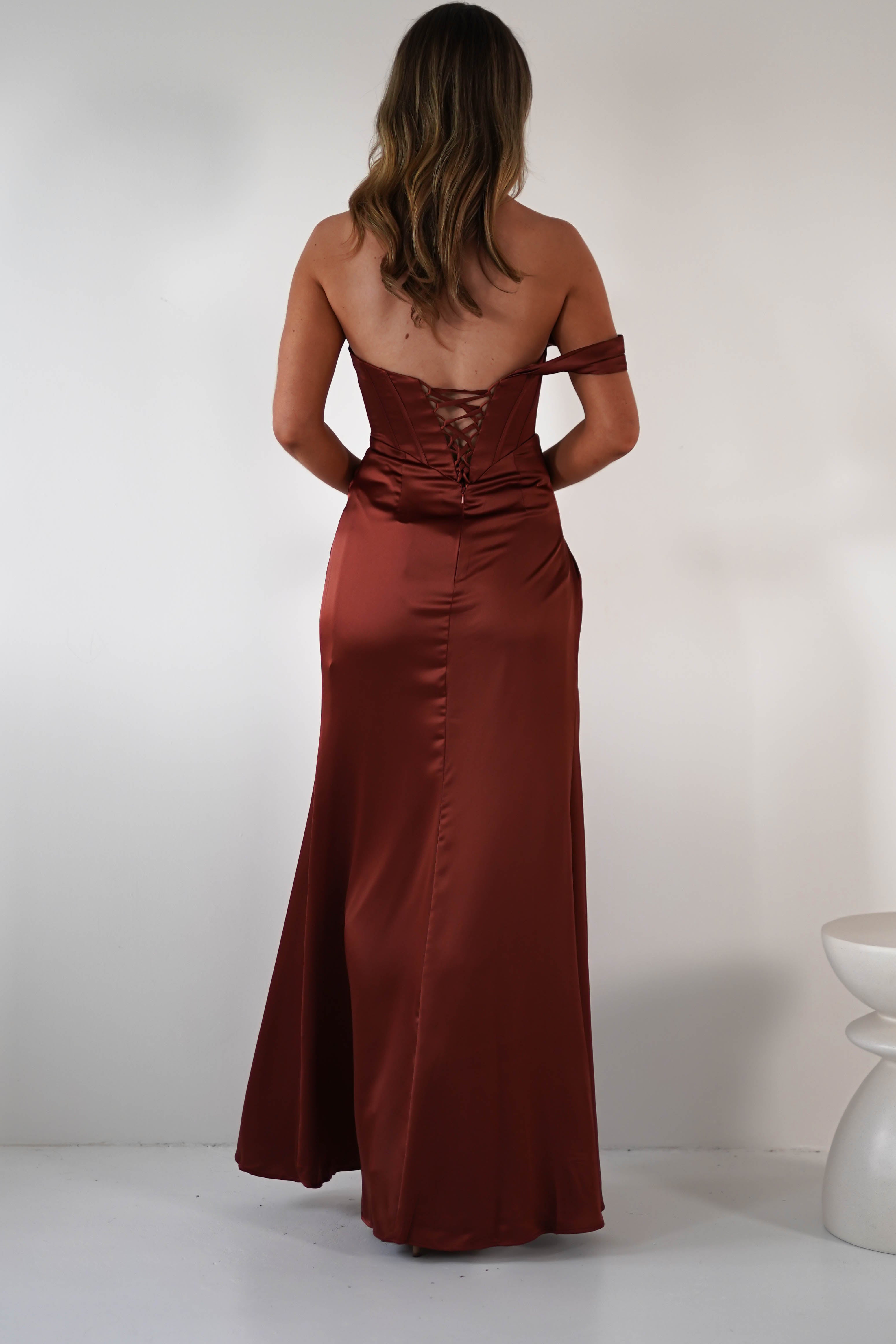 A woman with wavy hair stands facing away, wearing a floor-length, rust-colored satin dress with padded cups and a lace-up back. The off-the-shoulder gown features a high side slit, in a minimalist room with white walls and a modern white stool.