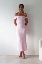 A woman stands against a white wall, smiling in a light pink, off-the-shoulder midaxi  with a sash on one sleeve. With her hands behind her back and hair in a loose updo, she’s the perfect inspiration for an elegant wedding guest look.