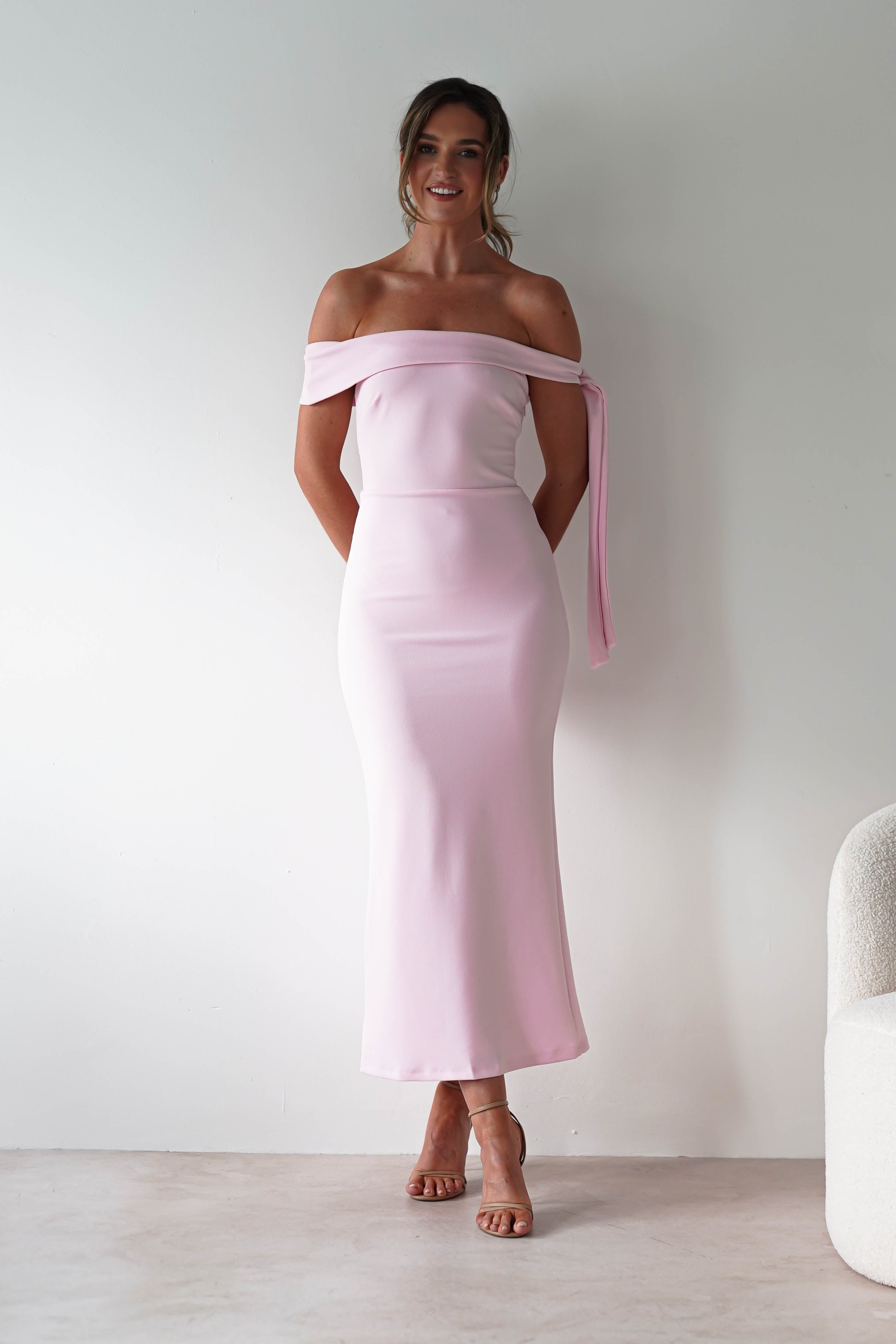 A woman stands against a white wall, smiling in a light pink, off-the-shoulder midaxi dress with a sash on one sleeve. With her hands behind her back and hair in a loose updo, she’s the perfect inspiration for an elegant wedding guest look.
