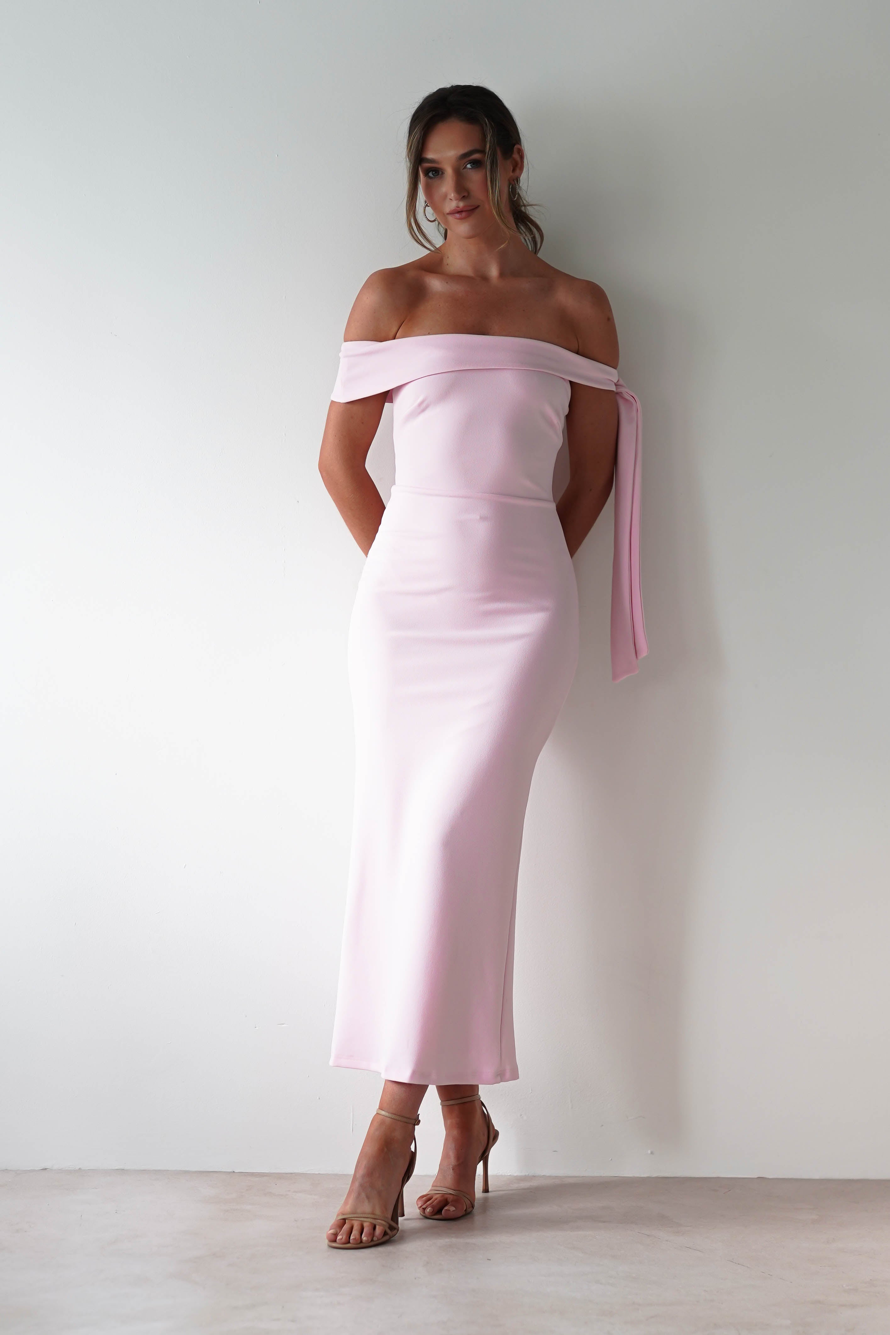 A woman stands against a white wall, wearing a fitted, off-the-shoulder, light pink midaxi dress with a tie detail on one arm and nude heeled sandals—an elegant look perfect for a wedding guest. Her hair is styled in a loose updo.