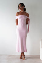 A woman stands against a plain light-colored wall, wearing an off-the-shoulder, fitted pastel pink midaxi  and nude heels. Her hair is styled in a loose updo, perfect for a wedding guest look, with her hands behind her back.