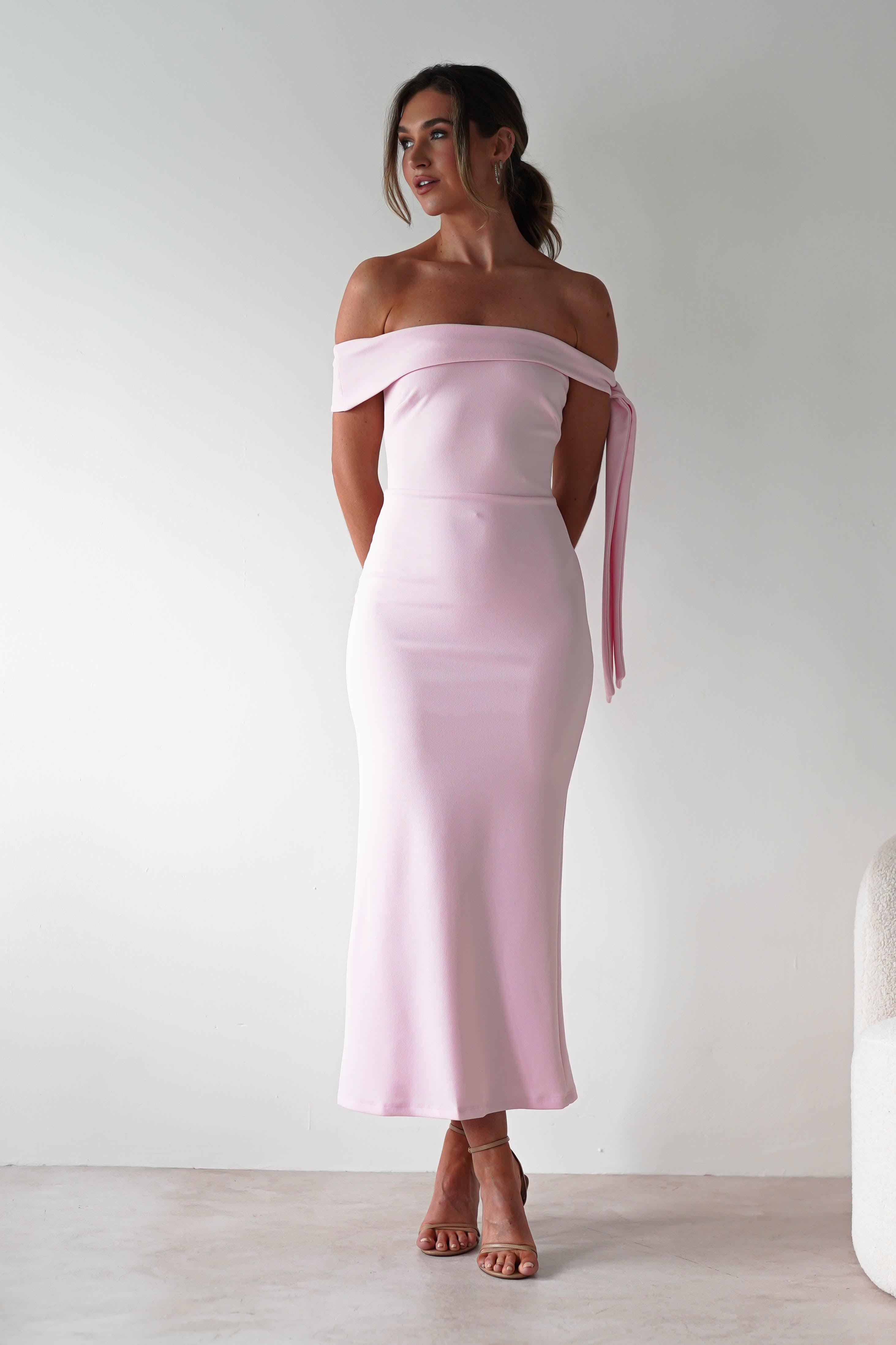 A woman stands against a plain light-colored wall, wearing an off-the-shoulder, fitted pastel pink midaxi dress and nude heels. Her hair is styled in a loose updo, perfect for a wedding guest look, with her hands behind her back.