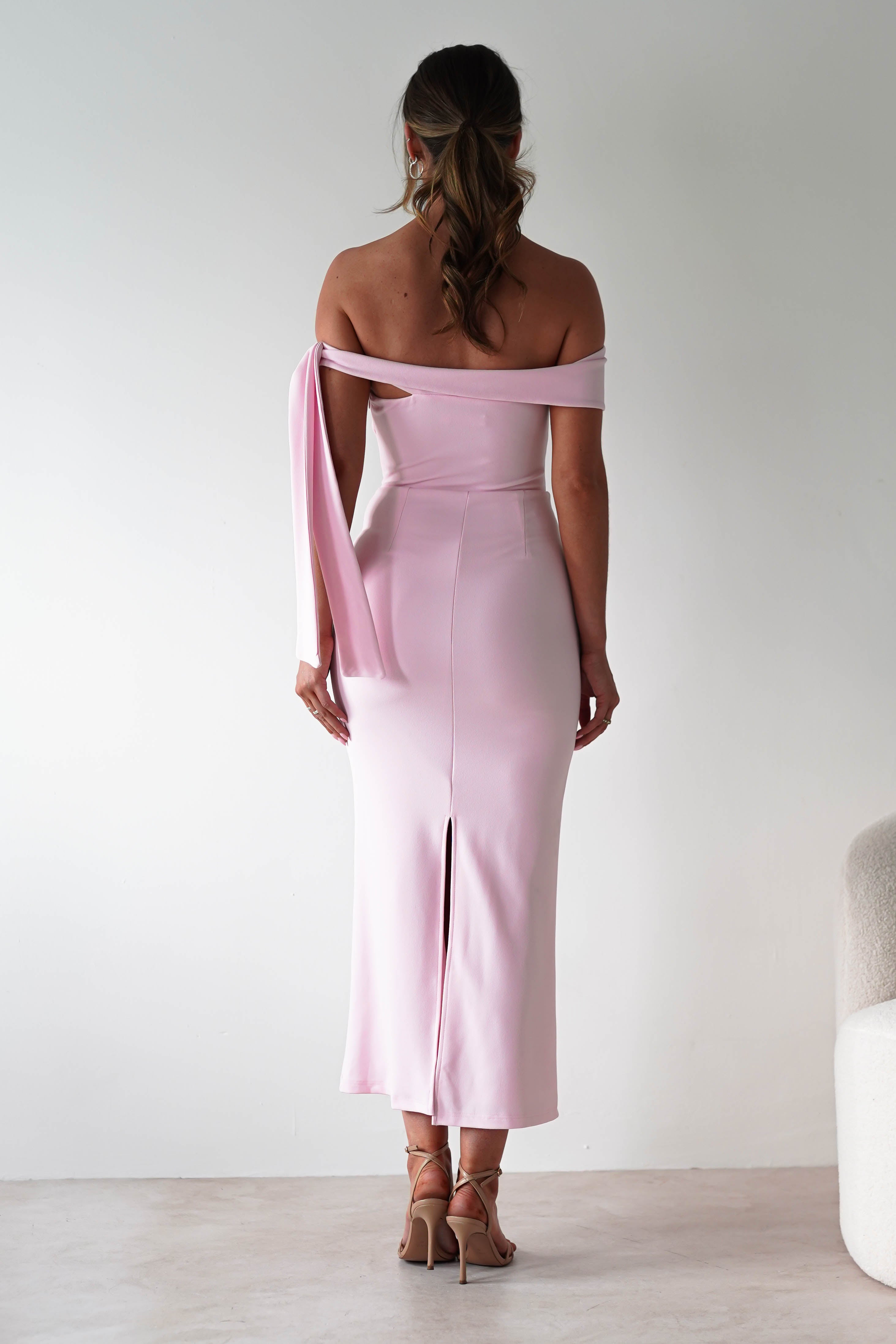 A woman stands facing away, wearing a fitted, off-the-shoulder midaxi dress in light pink with a back slit. Her hair falls in loose waves, complemented by nude high-heeled sandals—a chic choice for any wedding guest in a minimalist setting.