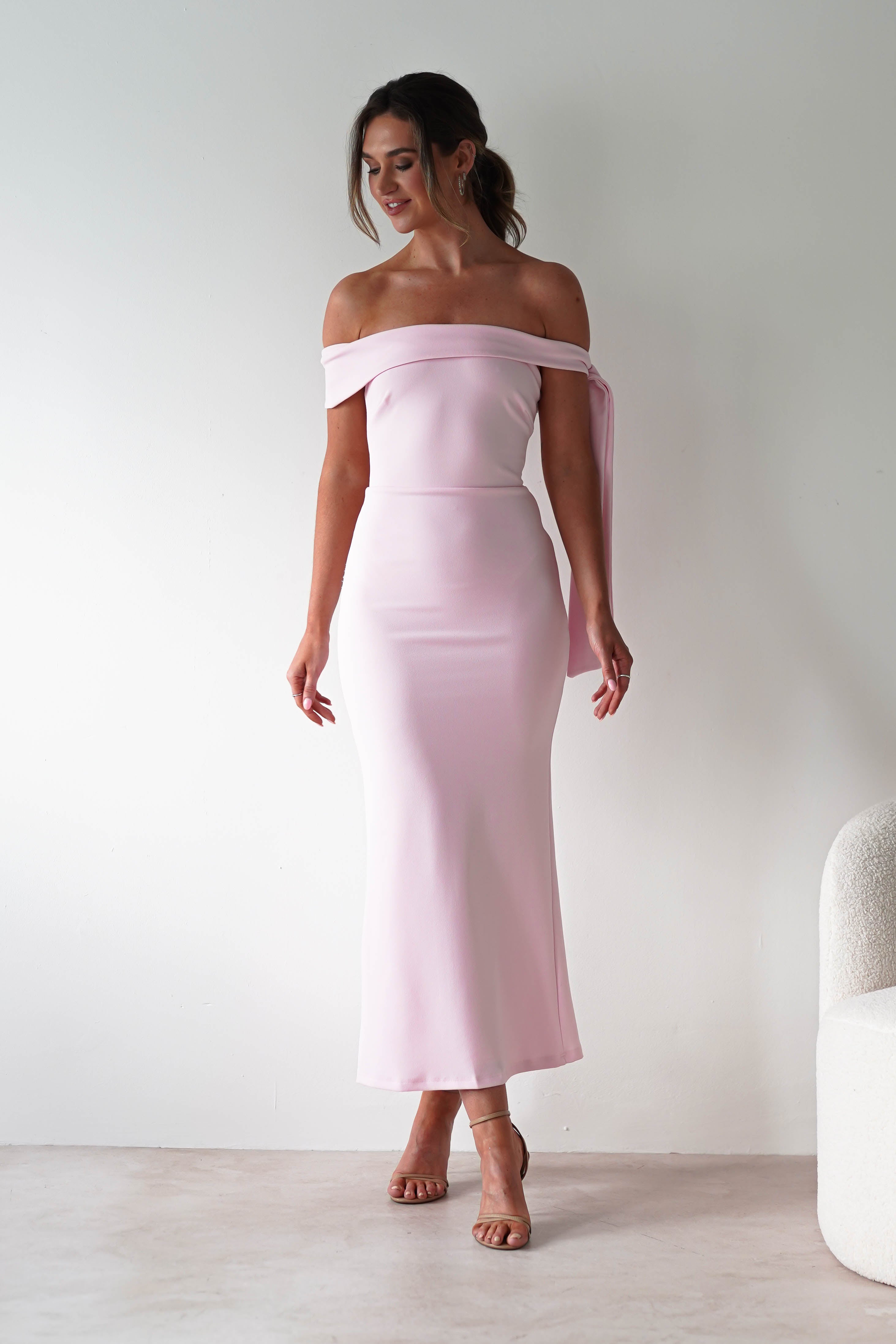 Woman wearing a light pink, off-the-shoulder midaxi dress and nude heels, standing indoors against a plain white wall and looking to the side, smiling—perfect for a stylish wedding guest look.