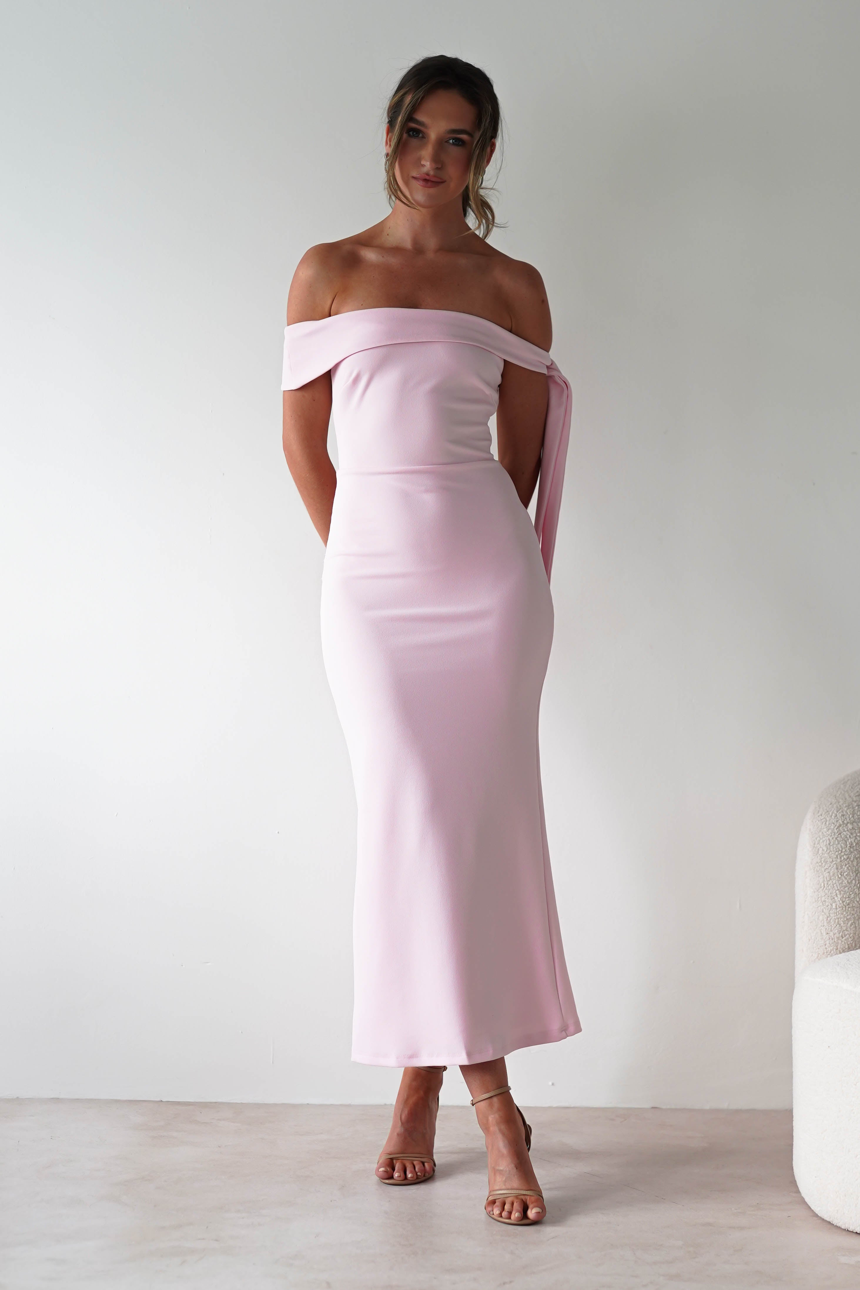 A woman stands against a plain white background wearing a light pink, off-the-shoulder midaxi dress with a fitted silhouette—an elegant choice for any wedding guest. Her hair is tied back and she completes the look with strappy high-heeled sandals.
