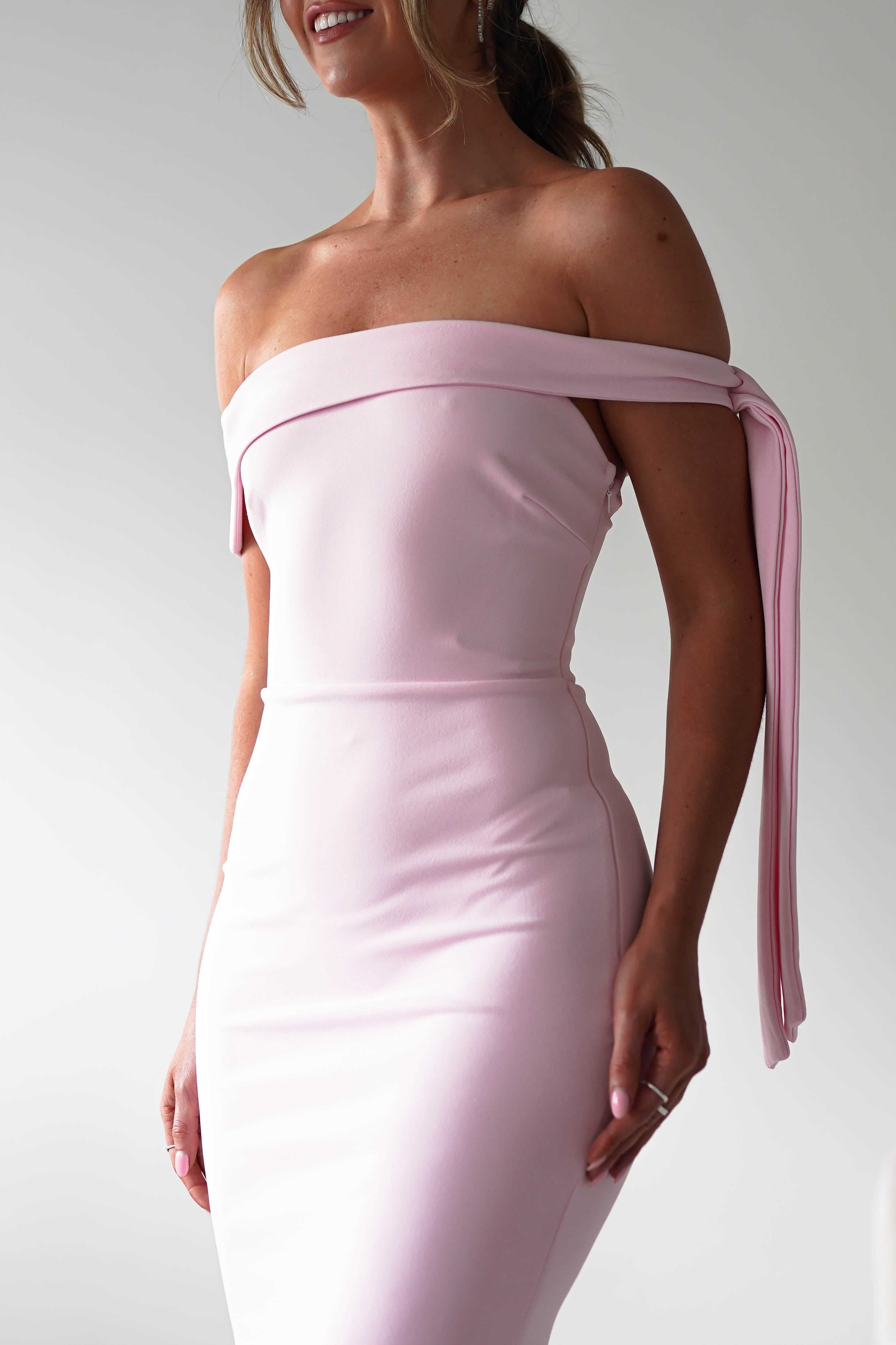Woman wearing a fitted, off-the-shoulder light pink midaxi dress with a decorative fabric tie on one arm, standing against a plain light background. Her head is partially out of frame, and she is smiling—a lovely choice for a wedding guest.