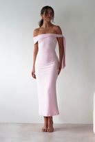 A woman stands against a plain white background, wearing a light pink, off-the-shoulder midaxi  with a sash detail on one arm—perfect for a stylish wedding guest. Her hair is tied back and she finishes the look with nude heels.