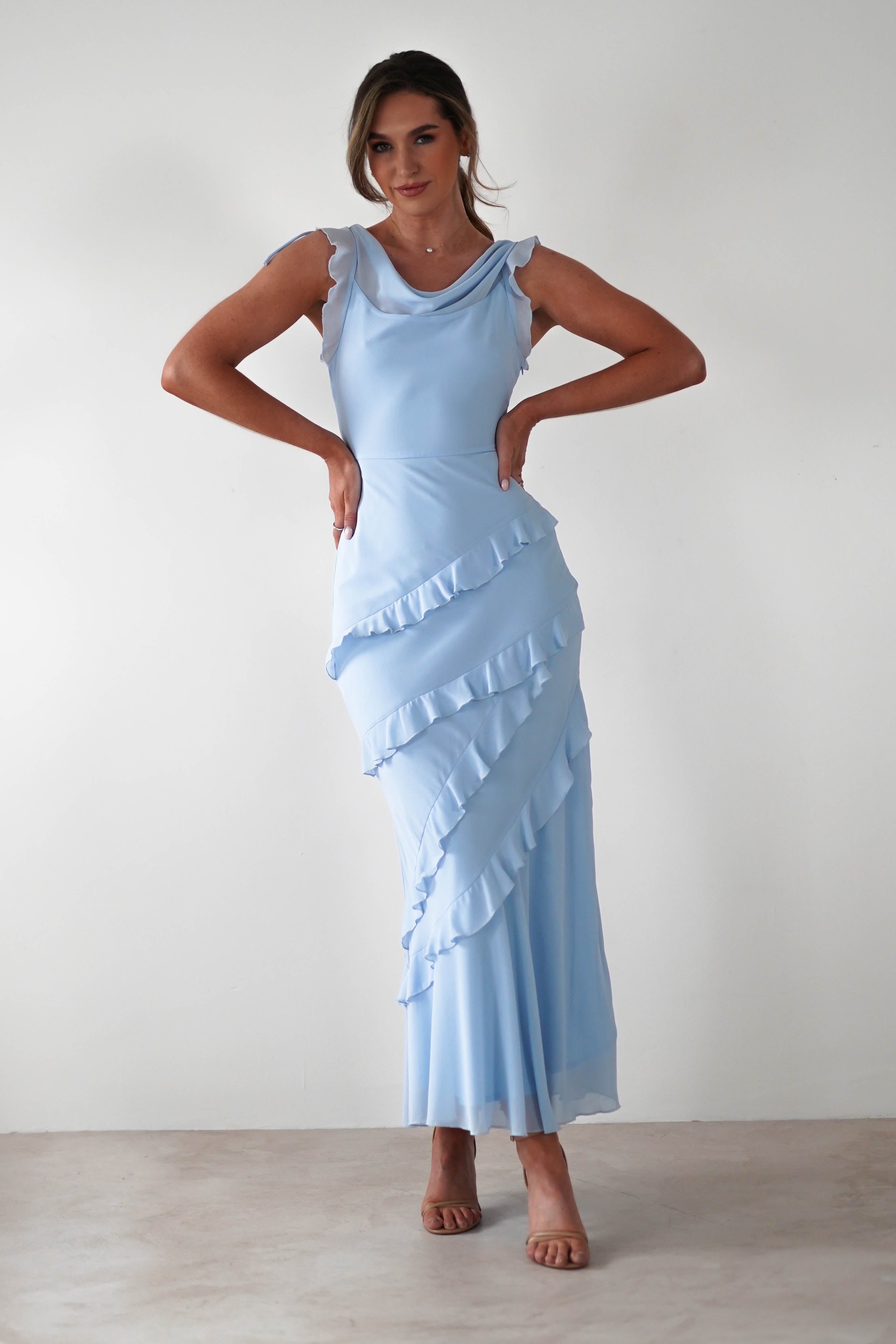 A person stands against a white background, exuding elegance in a light blue satin sleeveless dress with ruffled layers, perfect for special occasions. They pose confidently with hands on hips and wear brown heels, while their hair is styled up.