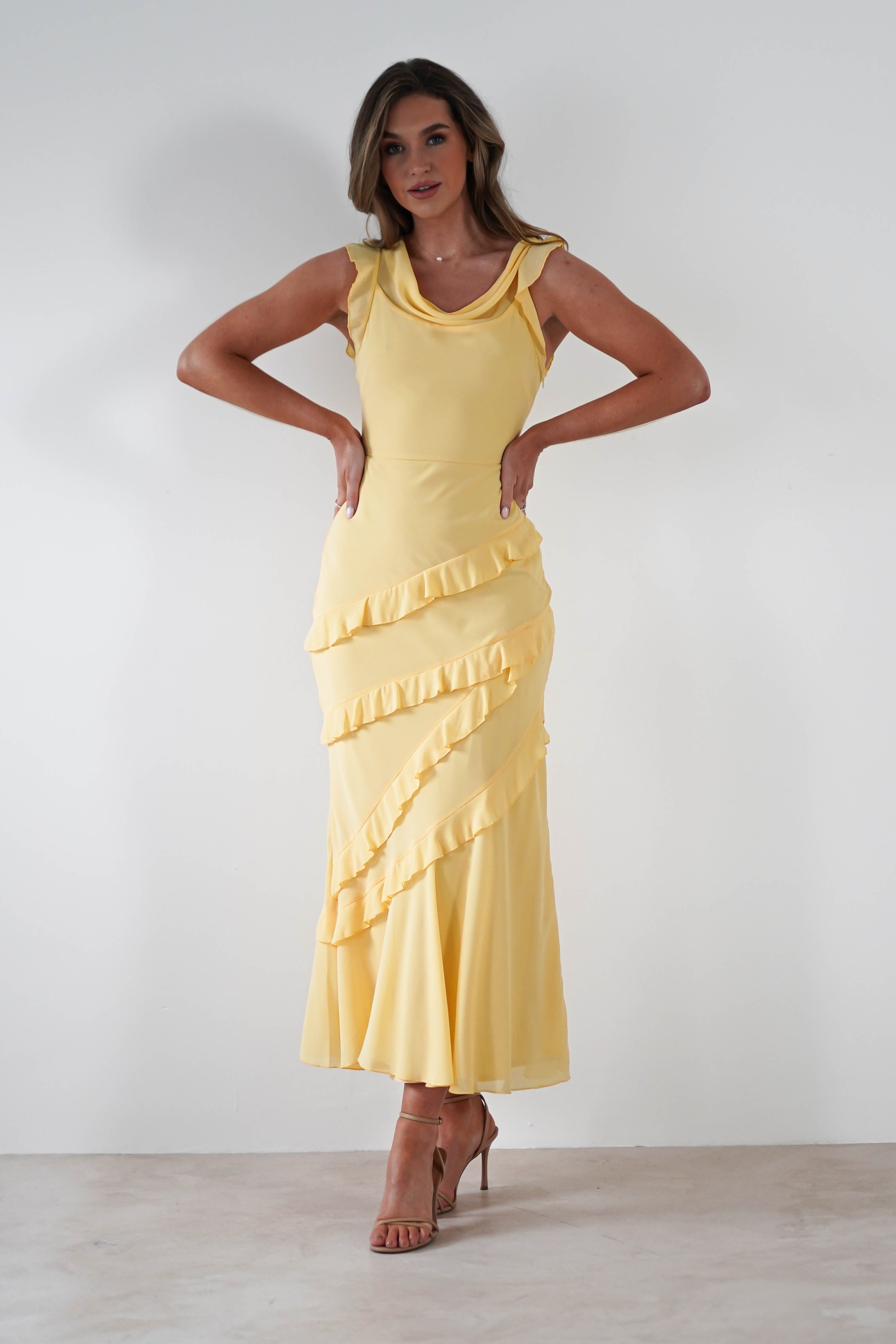 A woman stands against a plain white background wearing a yellow maxi dress with ruffled diagonal details in soft knit satin and brown heeled sandals. Her hands are on her hips as she looks confidently at the camera—perfect for special occasions.