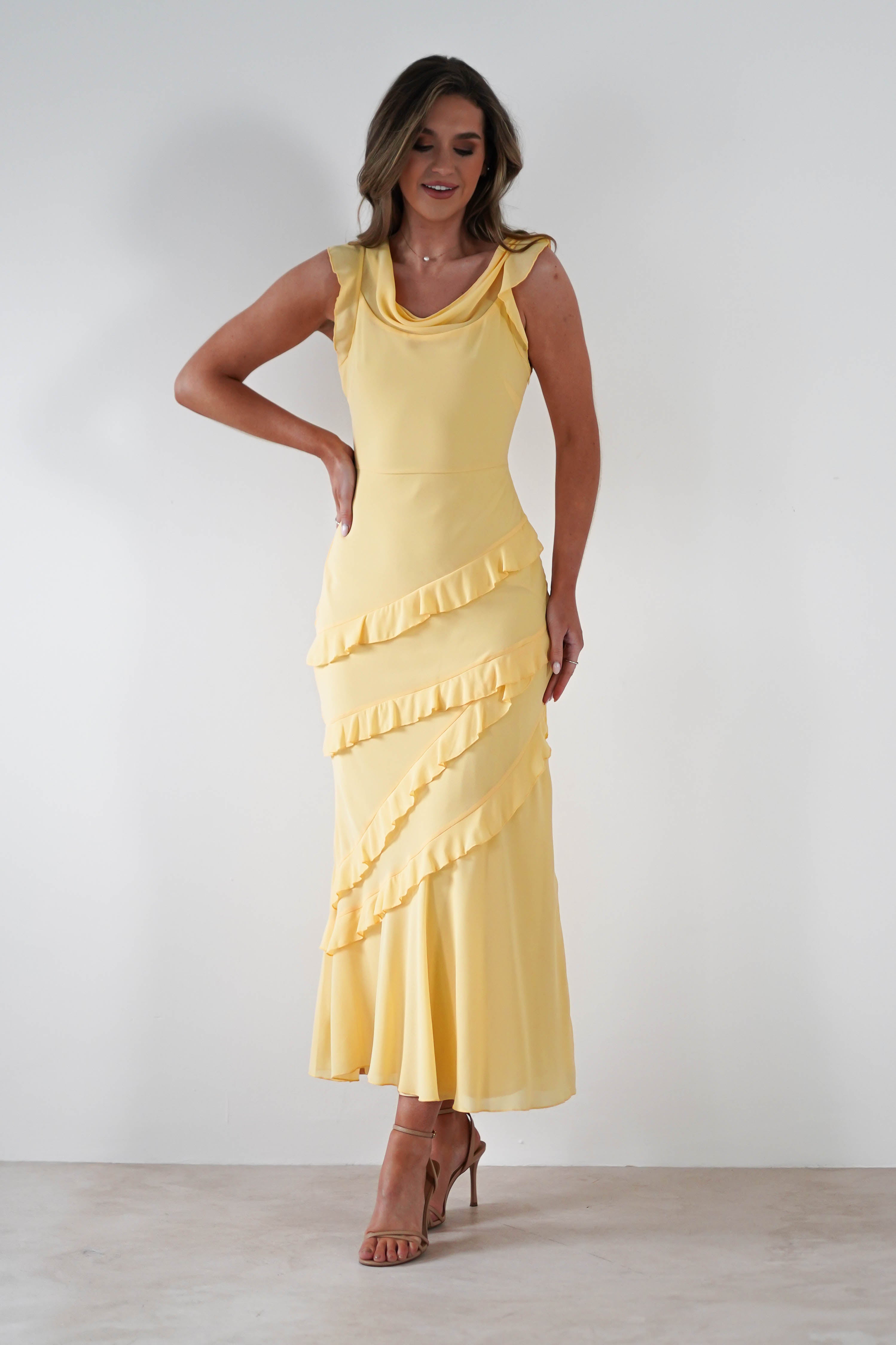 A woman in a sleeveless, floor-length yellow maxi dress with diagonal ruffle details stands against a plain white background, posing with one hand on her hip and wearing tan high-heeled sandals—perfect for special occasions.