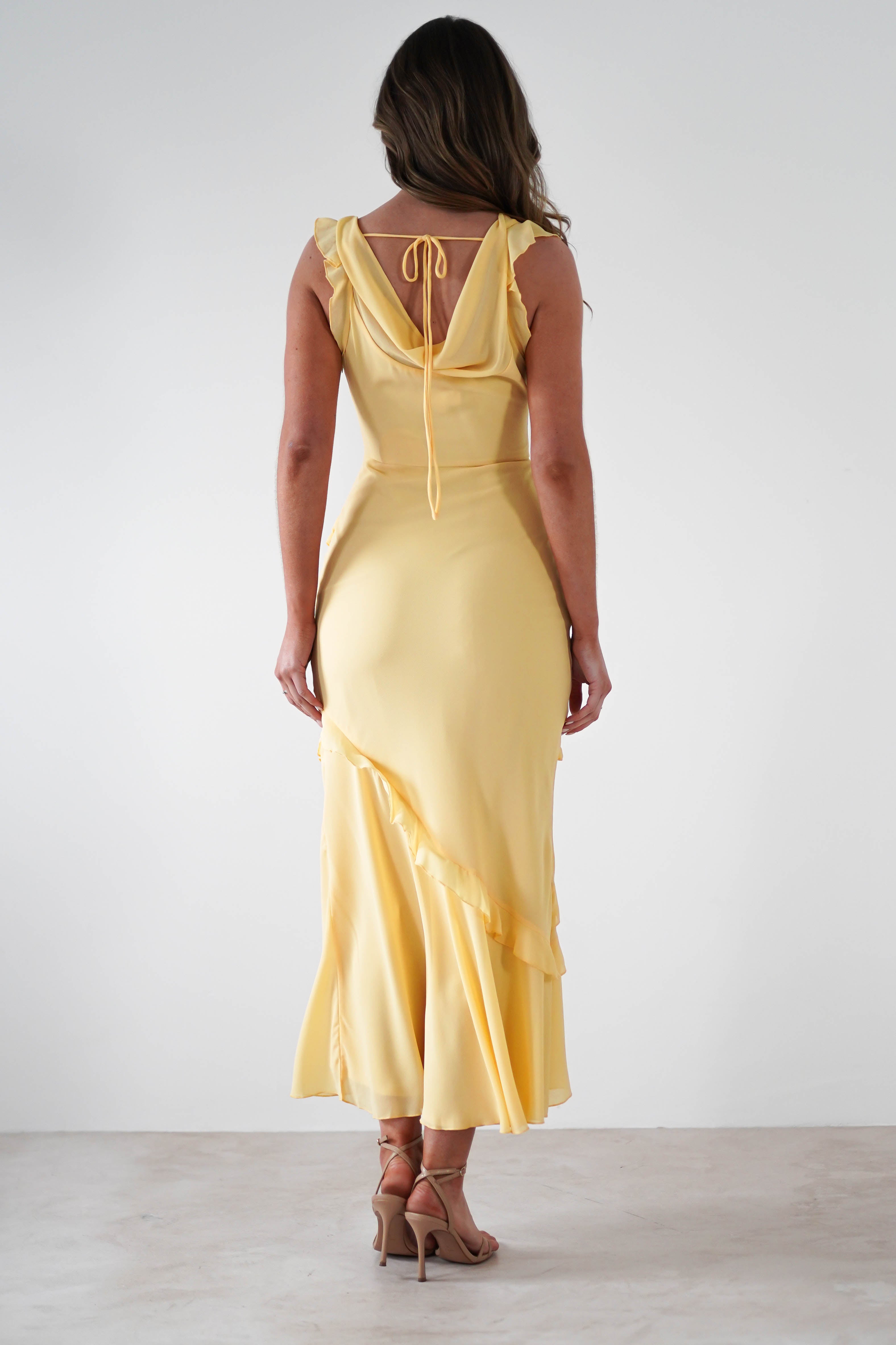 A woman stands facing away, wearing a soft knit satin maxi dress in pale yellow with ruffled details and a tie at the upper back—an elegant choice for special occasions. She has wavy brown hair and nude high-heeled sandals against a plain white background.