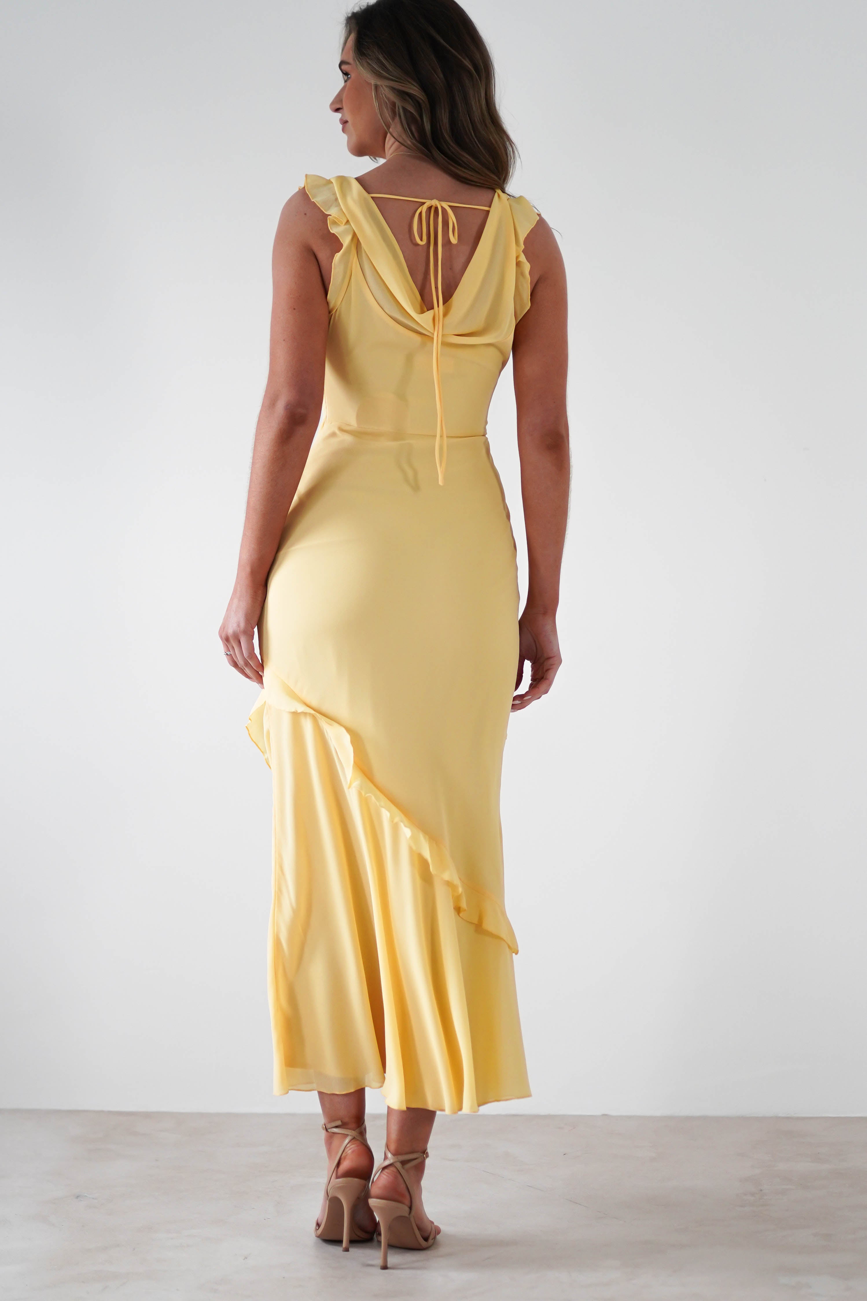 A woman wearing a light yellow satin fabric maxi dress with ruffle details and tie-back stands facing away, showing the back of the dress and beige high-heeled sandals—perfect for special occasions—against a plain background.