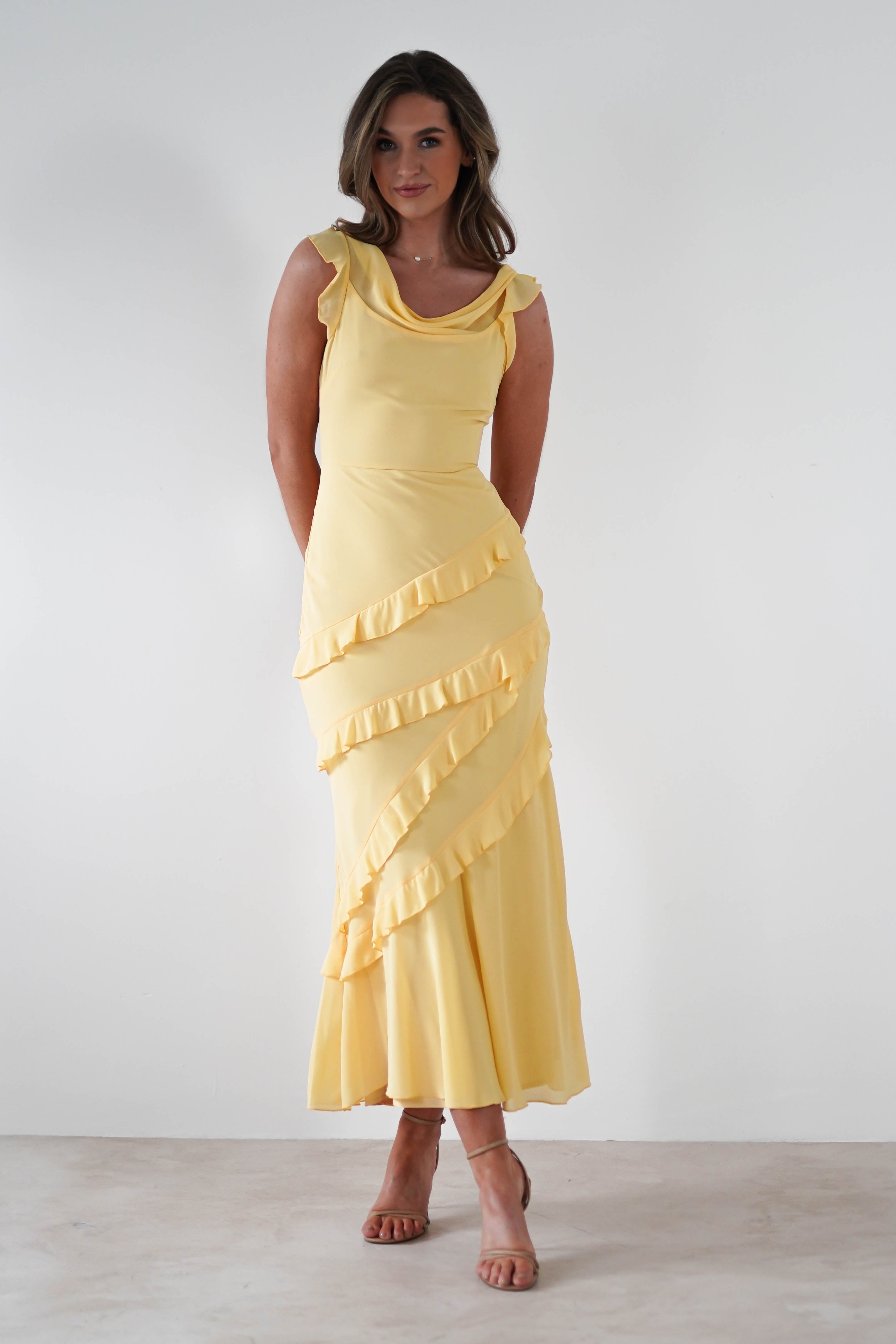 Woman wearing a long, sleeveless yellow maxi dress with diagonal ruffle details in soft knit satin, perfect for special occasions, standing against a plain white background.