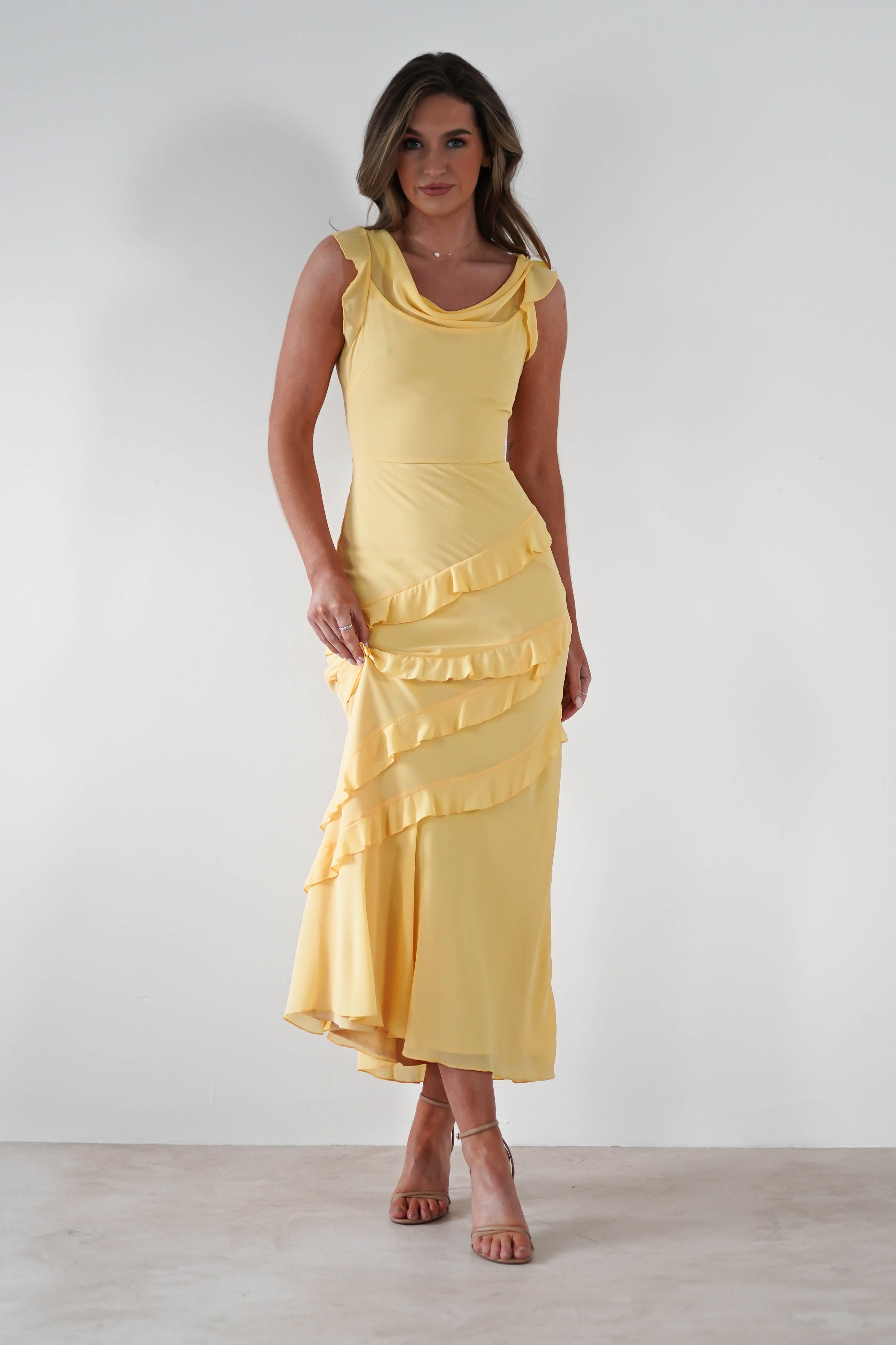 A woman wearing a sleeveless, floor-length yellow maxi dress with ruffle details poses against a plain white background. She has long hair and is standing in strappy heels, perfect for special occasions.