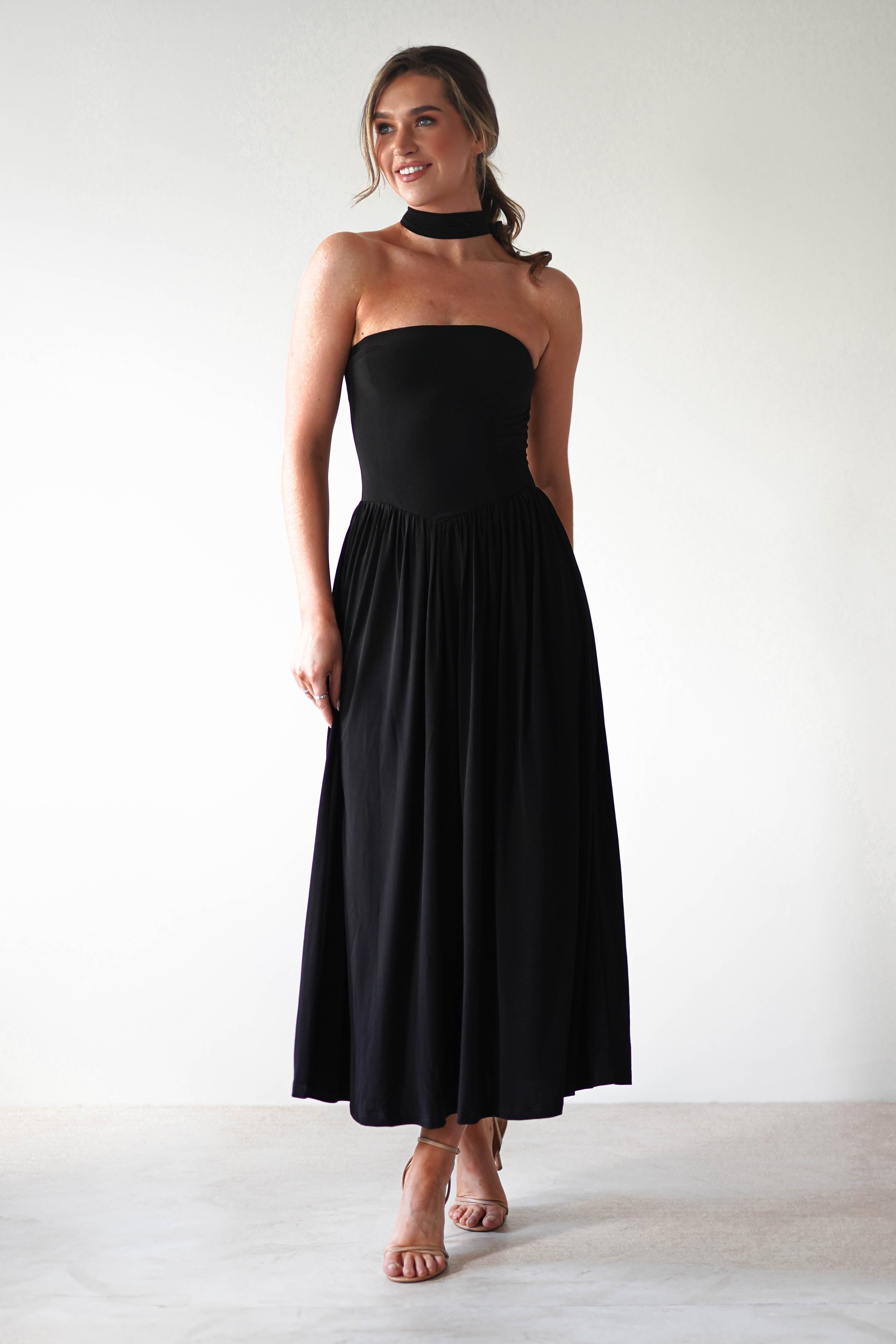 A woman stands against a plain light background, wearing a strapless black midi dress with a pleated skirt and matching black choker—an elegant choice for a wedding guest. She has light skin, brown hair styled back, and wears nude heeled sandals.